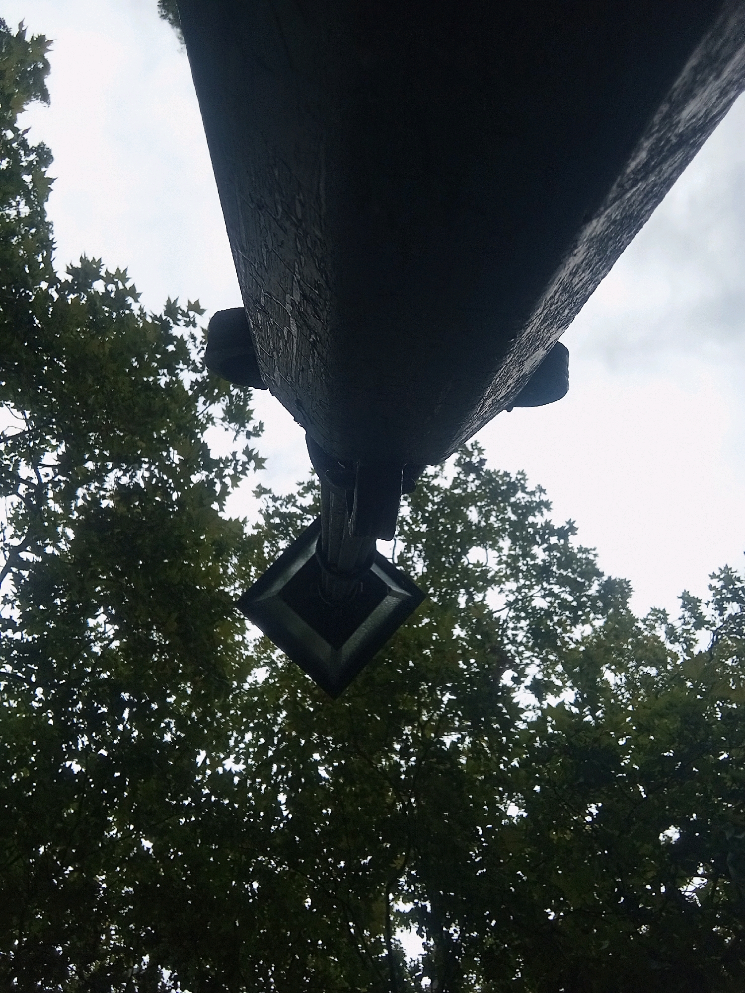 This low-angle, close-up photograph captures a portion of a large, dark, weathered wooden beam against a bright, overcast sky. The beam has visible texture and appears to be part of a larger structure, possibly an overhang or a very thick post. Hanging from the beam is a dark, square-shaped object with a decorative element at its base, resembling a lantern or a decorative finial.
The background is filled with the dense foliage of green trees, suggesting an outdoor setting. The leaves are large and appear to be from sycamore trees, based on their characteristic shape. Light filters through the canopy, creating a dappled effect. The overall impression is of a view looking upwards through trees towards a slightly ominous sky, with a man-made object suspended overhead.
This image does not depict any people, actions, or interactions. The scene is static and focuses on the architectural and natural elements.
This image was taken in Baden, Austria. The specific location within Baden is the historic theatre building (Stadttheater Baden). The dark beam and decorative fixture are part of the exterior architectural ornamentation of this building, specifically the ornate carved wooden elements on the theatre's facade, which were created by Josef Daniel von Hubner in the late 19th century. The view is from below, looking up at these decorative details and the surrounding trees.