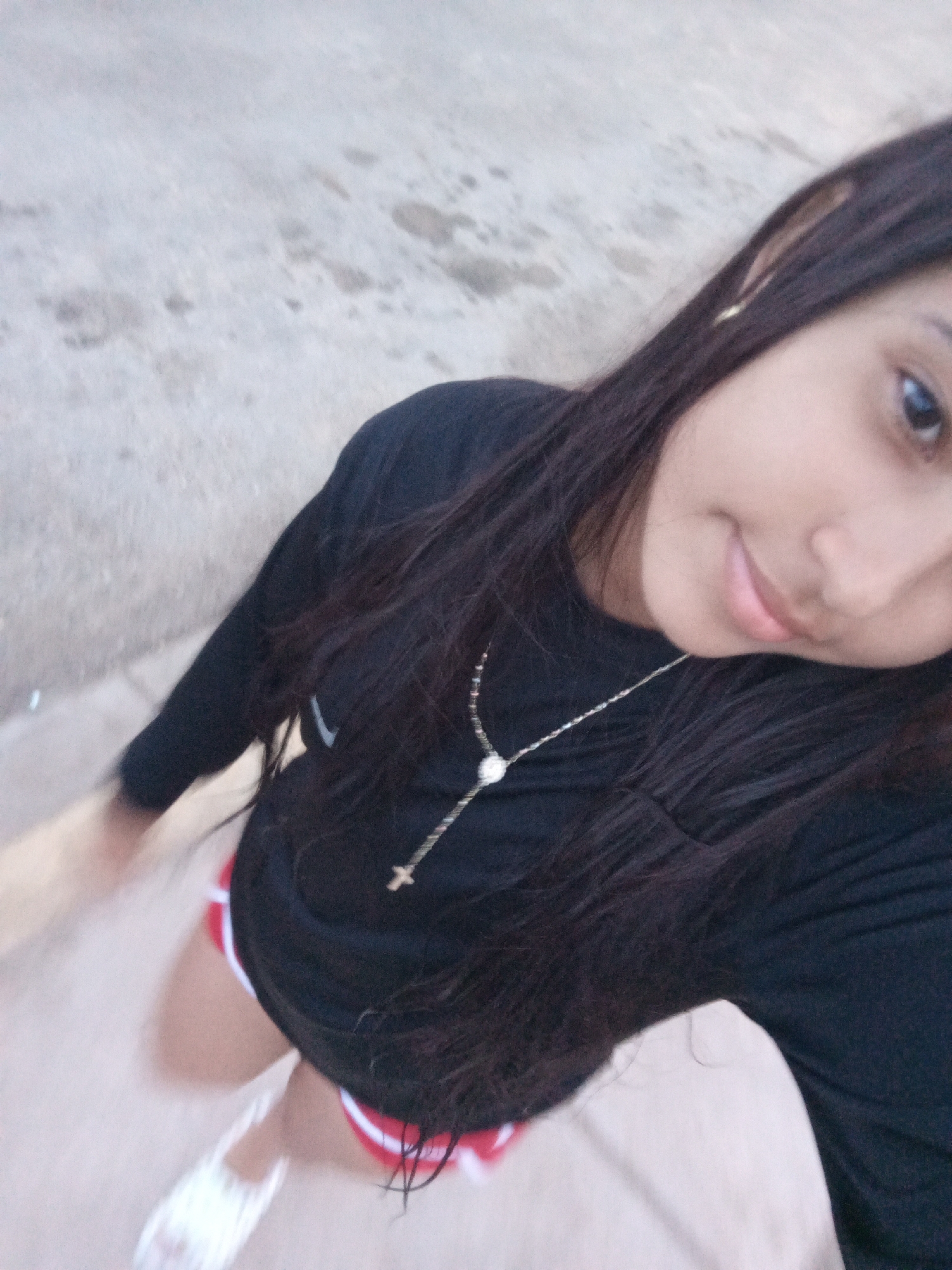 A young woman with long, dark hair is taking a selfie outdoors. She is wearing a black long-sleeved shirt and a silver necklace with a cross pendant. Her shorts are red with white trim, and she appears to be wearing white sandals. The background is a light-colored concrete surface. The image does not provide enough details to confirm that the location is Juan Griego, Venezuela.