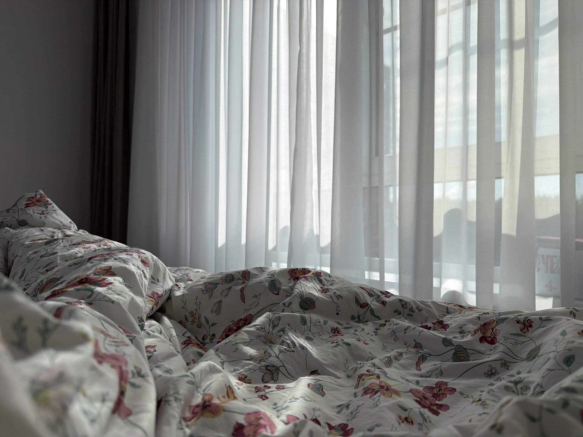 The image depicts a close-up view of a rumpled bed with floral patterned sheets, facing a window covered by sheer white curtains. The curtains are illuminated by sunlight, creating a soft, diffused light that casts shadows on the bedding. To the left of the curtains, a darker, opaque curtain is visible. The scene appears to be indoors, likely a bedroom. There are no people or animals present. The environment suggests a domestic setting, potentially in a city like Drozhzhino, Russia, given the context. The focus is on the textures of the fabric and the play of light and shadow.