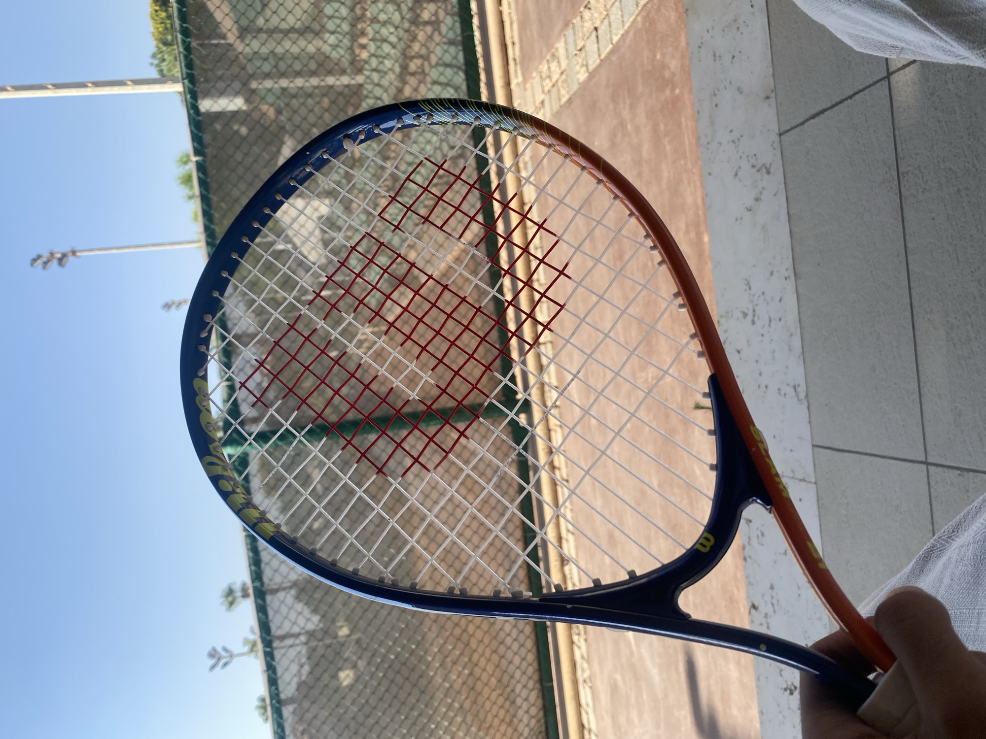 A close-up shot captures a tennis racket against a backdrop of a tennis court and a bright blue sky. The racket, held by a person whose hand is partially visible at the bottom left, features a blue and orange frame. Its strings are white, with a pattern of red strings forming a heart shape in the center. The court's green fence and lights are visible behind the racket. The foreground shows light-colored tiled flooring. The scene is likely situated outdoors in Sheikh Zayed, Egypt, given the architecture and style of the court and surrounding facilities.