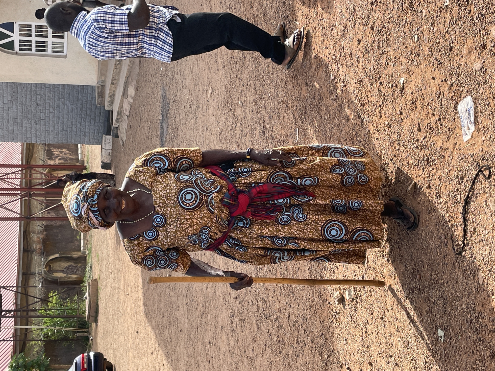 The image shows an elderly woman standing on a dirt ground, using a wooden walking stick. She is wearing a patterned brown and blue dress and a matching head wrap, along with a white pearl necklace. A red sash is tied around her waist. A man in a checkered shirt and black pants is visible in the background. To the left, there is a building with a red roof, scaffolding, and some greenery. In the far background, there is a grey brick building and a white building with a window. The location appears to be outdoors with a ground covered in small rocks and dirt. The picture was taken in the city of Jos, Nigeria.
