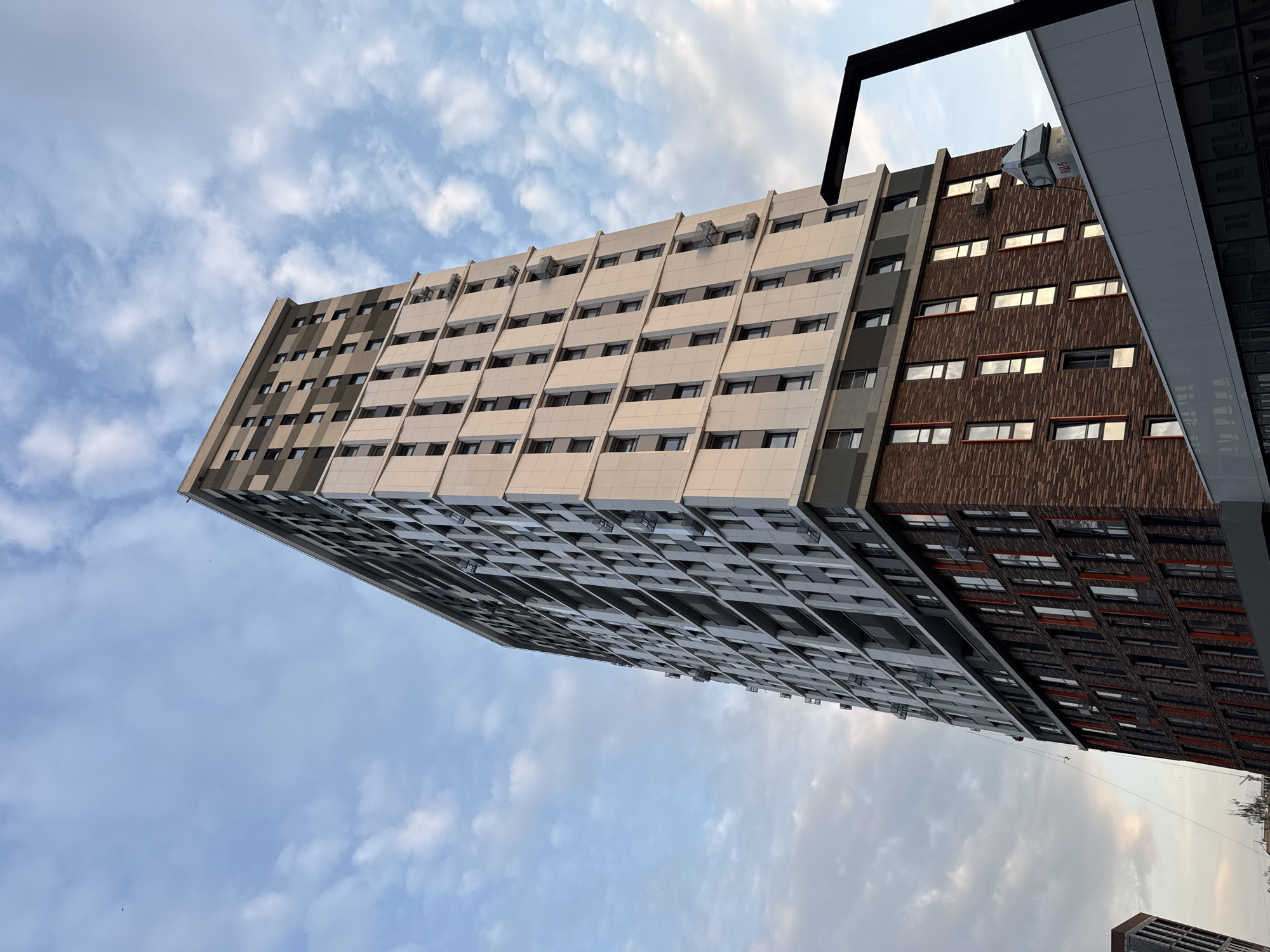 The image shows a modern, multi-story residential building under a cloudy sky. The building features a facade of light-colored panels and dark grey sections, with a lower portion clad in brickwork featuring vertical red accents. Windows are visible throughout the structure. A black, angular lamppost is partially visible in the foreground on the right. The photograph is taken from a low angle, looking upwards at the building, with the sky occupying a significant portion of the frame. This building is identified as the "Grand Park" residential complex, located in Izhevsk, Russia. There are no people clearly visible in the image.