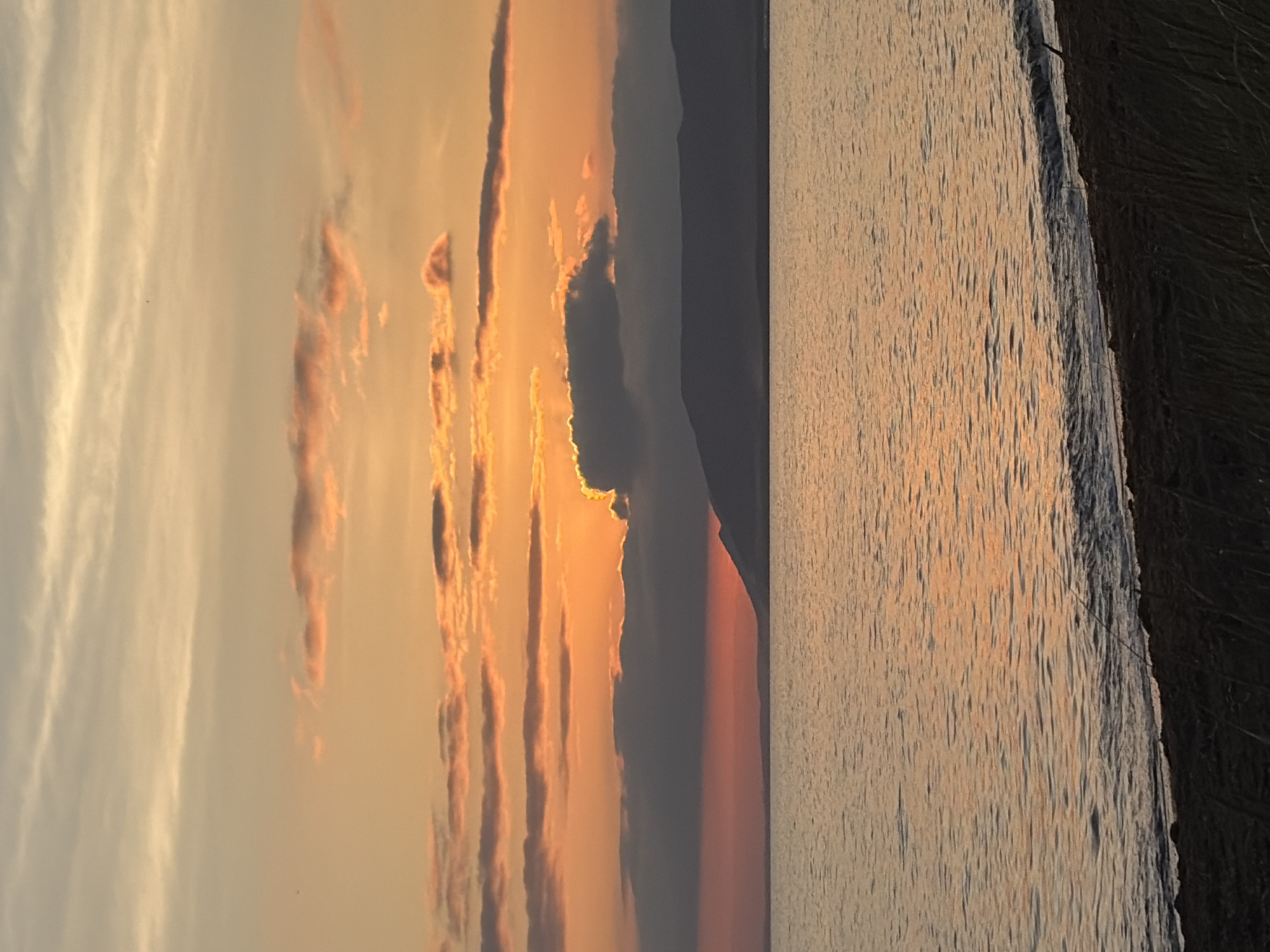 This image depicts a sunset over a body of water, with a silhouetted mountain range in the distance. The sky is filled with clouds painted in hues of orange, pink, and yellow, with some darker, gray clouds partially obscuring the sun. The water is calm and reflects the warm colors of the sky. A dark, sandy beach with some vegetation is visible in the foreground. The location is identified as Ozerskoe, Russia. There are no people or discernible actions in the image.