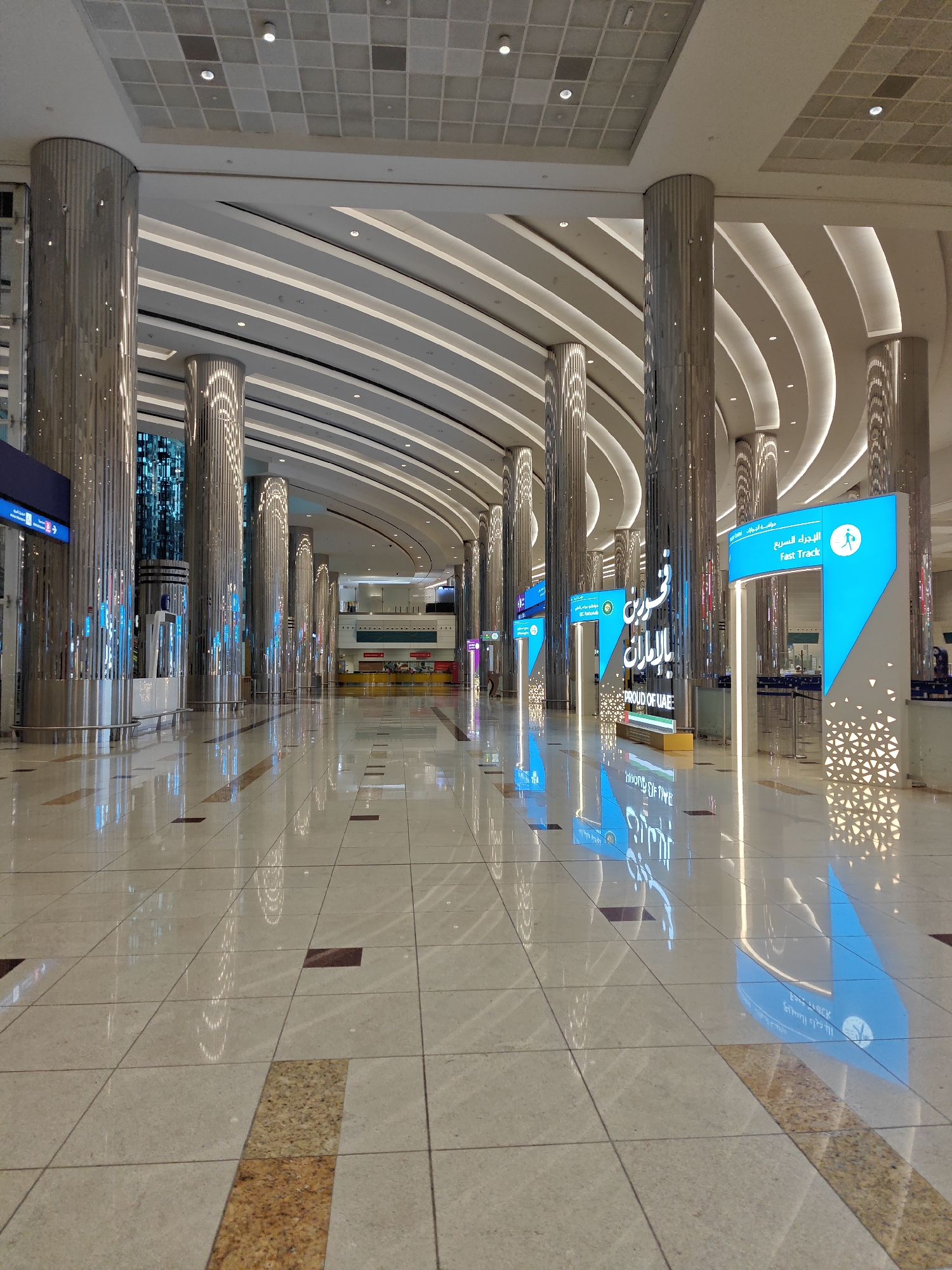 The image shows an interior view of a modern airport terminal in Dubai, United Arab Emirates. The hall features a highly polished marble floor with reflective qualities, catching the light from the illuminated ceiling and the surrounding signage. Large, cylindrical, polished metal columns line the walkway, adding to the sense of grandeur and spaciousness. The ceiling is a complex architectural design with curving, illuminated panels creating a dynamic visual effect. In the foreground, a walkway extends into the distance, flanked by the prominent columns.
Various signs are visible, some illuminated in bright blue and featuring Arabic and English text, indicating directions or services within the airport. One prominent sign reads "Fast Track" with a silhouette of a person walking. Another sign says "Proud of UAE." The overall atmosphere is clean, bright, and futuristic, characteristic of a major international airport. There are no people clearly visible in the image, suggesting the area is either uncrowded or the photo was taken at a time of low foot traffic. The environment appears to be an arrival or departure hall, given the presence of directional signage typical of such areas. The reflective floor and the clean lines of the architecture create a visually striking and contemporary space.