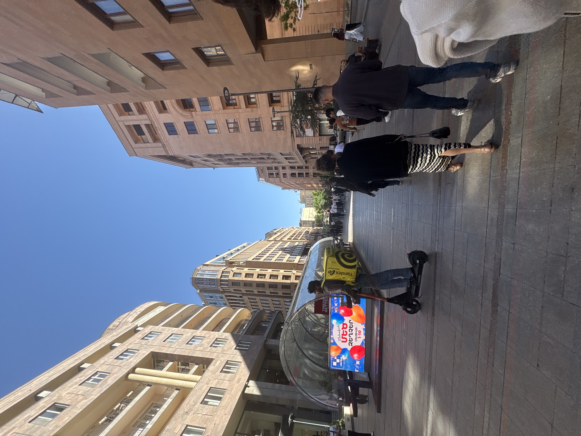 The image is a street-level view of a cityscape in Yerevan, Armenia. The sky is clear and blue. Buildings line both sides of the street, featuring light-colored facades with multiple windows and architectural details. Pedestrians are walking on the sidewalk. A delivery person with a yellow Yandex Go bag is riding an electric scooter. There is a curved glass-enclosed structure with a digital display showing an advertisement. The street is paved with light grey tiles.