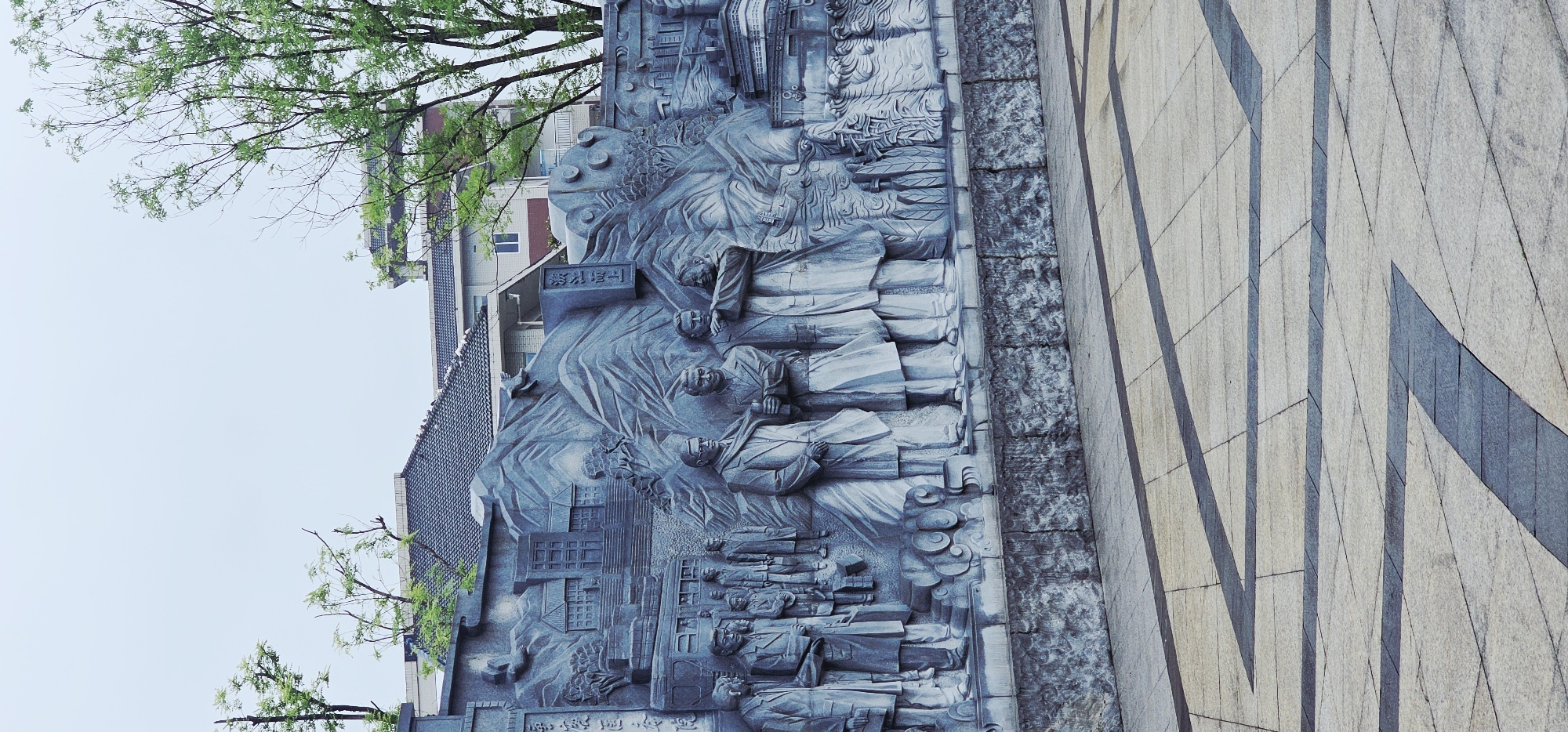 This is a full-shot, eye-level, outdoor, vertical-format, color photograph of a bas-relief sculpture in the Chongqing High-tech Zone, China. The sculpture, rendered in shades of grey stone, depicts a group of figures in historical attire, standing in front of a stylized mountainous landscape with buildings and a train. The figures are arranged in a frieze-like composition, with some individuals appearing to be in conversation or contemplation. Behind the figures, the sculpted landscape includes distinct mountain peaks and what appears to be a representation of traditional Chinese architecture. The sculpture is set against a muted sky with a few distant trees visible. In the foreground, a paved plaza with a geometric pattern of light and dark grey tiles leads up to the base of the sculpture. The overall impression is one of historical commemoration and artistic representation of a significant event or period.