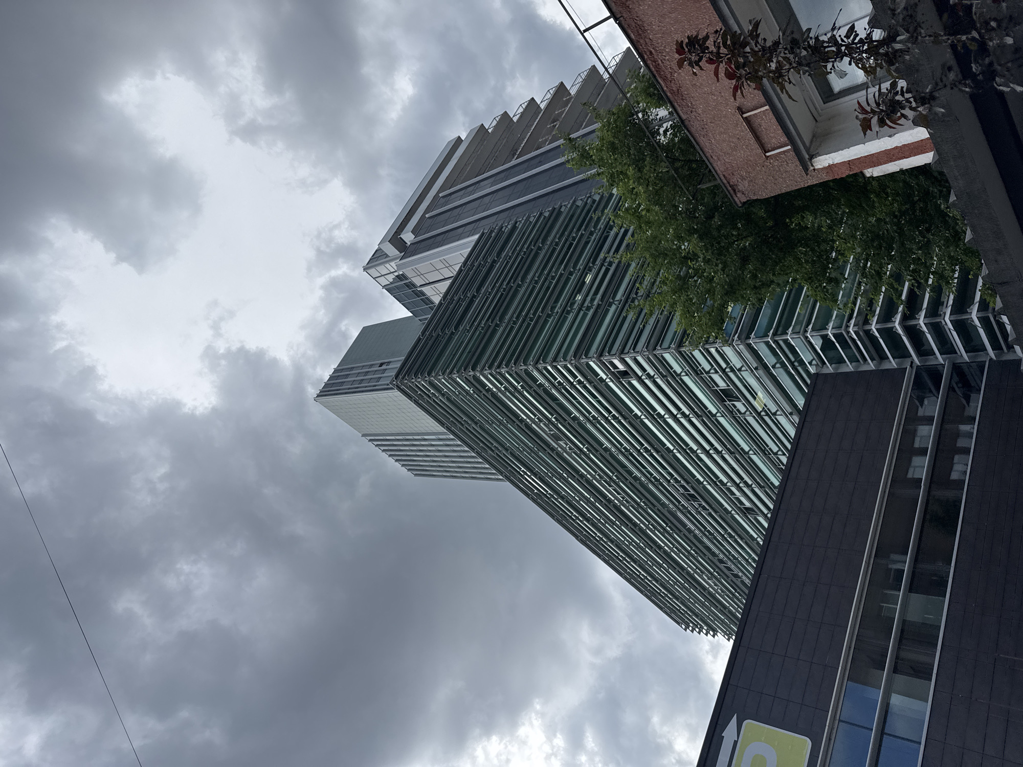 A low-angle, wide shot captures a cluster of modern buildings against a cloudy sky. The buildings are characterized by sleek architectural designs, with prominent use of glass and metal. One building features a facade composed of horizontal glass louvers, giving it a textured, layered appearance. Another adjacent building showcases a stepped design with balconies and dark vertical accents. A darker, lower building with reflective windows is visible in the foreground, partially obscuring the base of the taller structures. Lush green trees are interspersed between the buildings, providing a touch of nature amidst the urban landscape. The sky is filled with heavy, gray clouds, suggesting an overcast or impending stormy weather. There are no people visible in the frame.