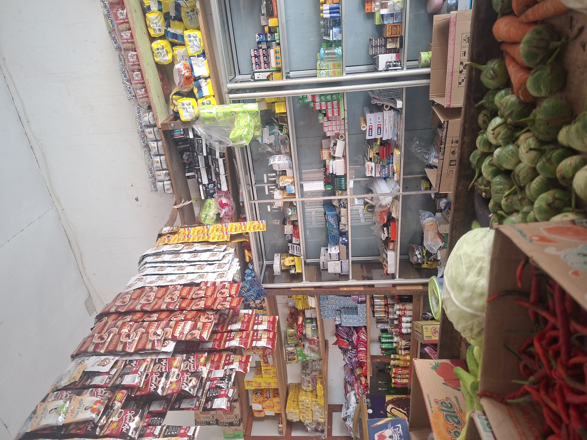 This is a medium shot, eye-level, indoor photograph of a small convenience store in Mulya Sari, Indonesia. The store is packed with goods displayed on wooden shelves and hung from the ceiling. The shelves are filled with packaged food items, drinks, and household supplies. In the foreground, a variety of fresh produce, including red chili peppers, green chilies, a head of cabbage, eggplants, and carrots, are arranged in cardboard boxes. The overall impression is one of a well-stocked, albeit somewhat cluttered, local market. There are no people visible in the image.