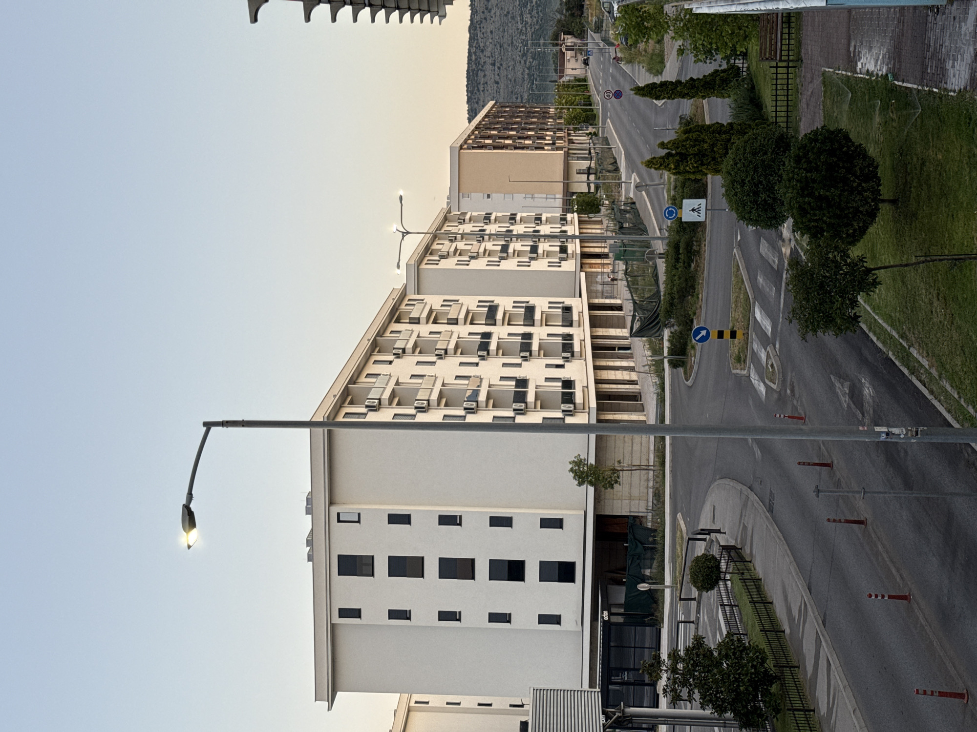 This is an outdoor, eye-level, vertical, medium shot of a street scene in a city, likely during dusk or dawn. The sky is clear and a pale blue. In the foreground, a street lamp with a lit bulb stands on the right side of the image, casting a soft glow. The street itself curves to the left, lined with modern, multi-story apartment buildings on the left and right. The buildings are primarily white and beige, with dark-framed windows and balconies. There are several trees and manicured bushes along the sidewalks and medians. Traffic cones and bollards are placed along the road. Traffic signs, including a speed limit sign (40) and a pedestrian crossing sign, are visible. The scene appears to be in a residential area with new construction. No people are visible in the image.