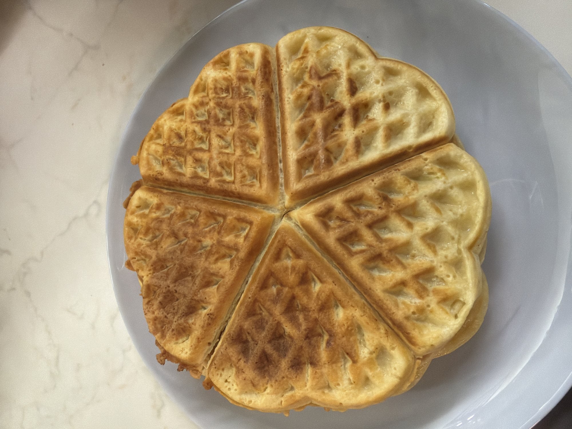 A top-down view shows a golden-brown waffle divided into five heart-shaped sections on a white plate. The waffle's surface has a grid pattern, and some sections appear darker than others. The plate sits on a white marble surface. There are no people, subjects, actions or interactions present in the scene. There are no indications of the location being Veliko Tarnovo, Bulgaria.