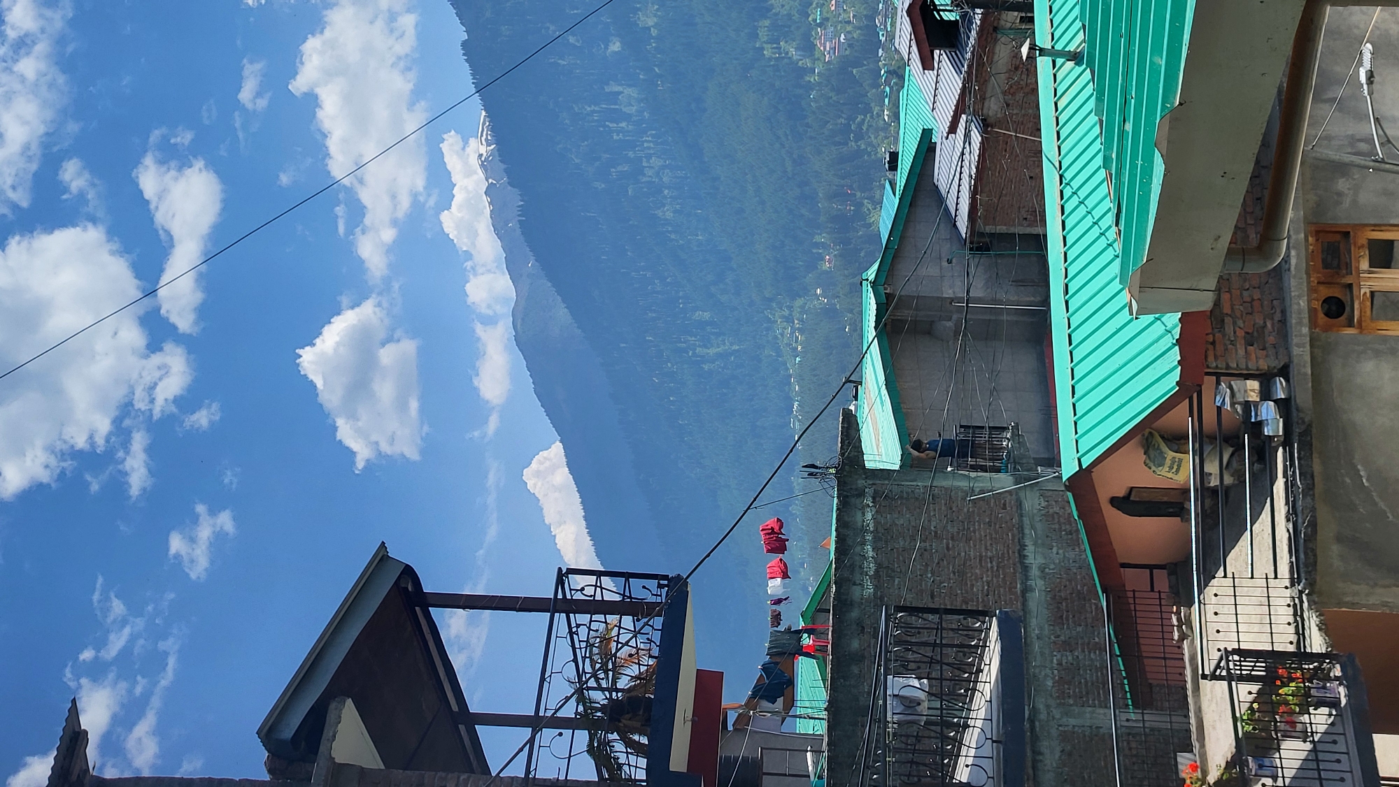 The image shows a view of rooftops and mountains in Vashisht, India. In the foreground, there are several buildings with teal-colored corrugated metal roofs. Balconies with metal railings are visible on some of the buildings. Laundry is hanging on a clothesline on one of the balconies. In the background, there are lush green mountains with some snow-capped peaks at the top. The sky is blue with scattered white clouds. The scene suggests a residential area in a mountainous region.