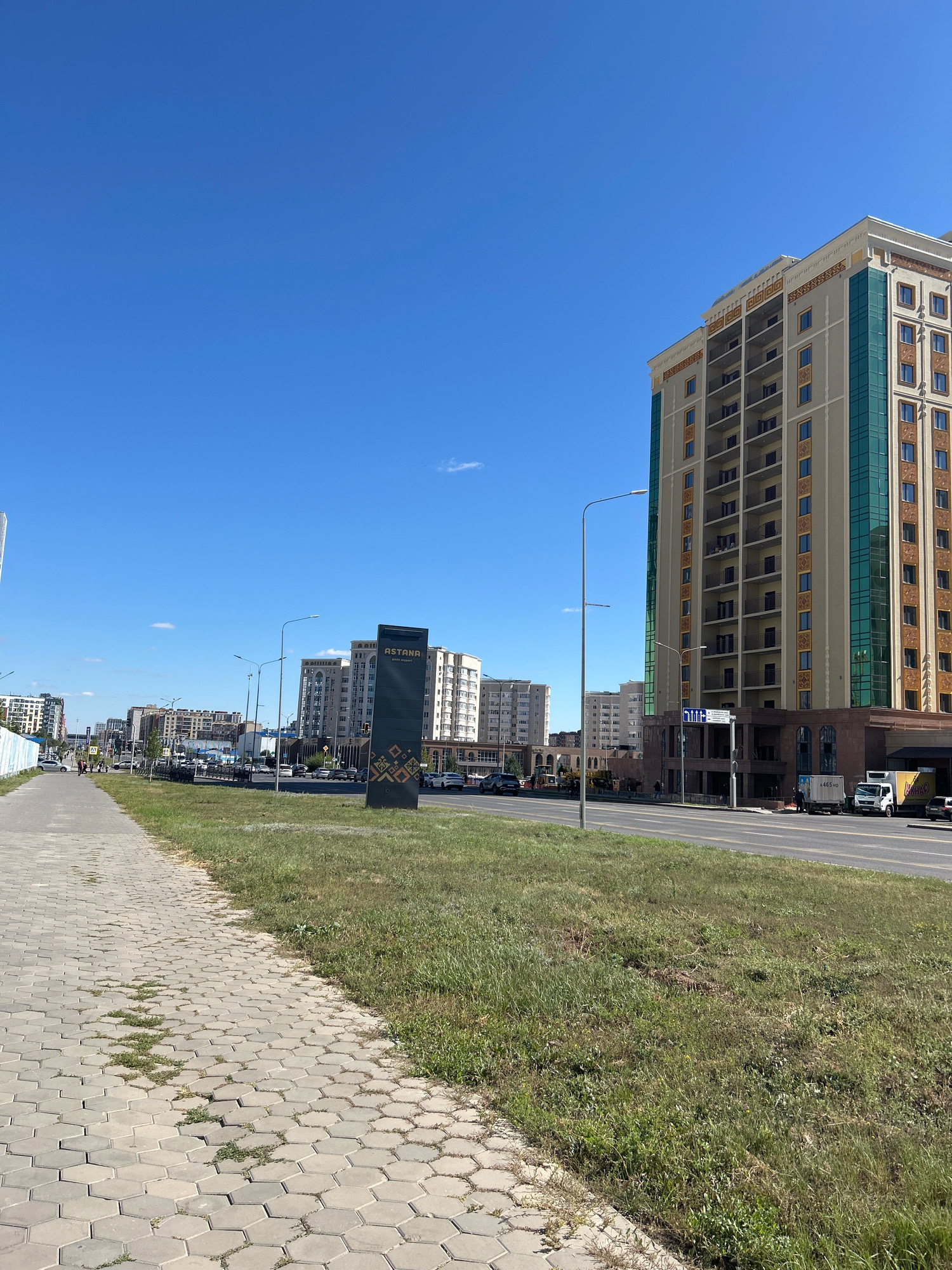 This is a street-level, eye-level, medium shot of a street in Astana, Kazakhstan. The scene is dominated by a bright blue sky with a few small white clouds. In the foreground, a paved sidewalk with hexagonal tiles runs along the left side of the image. The sidewalk is bordered by a grassy median with some patchy areas and weeds. A road with multiple lanes runs parallel to the sidewalk, with cars visible on it.
On the right side of the image, a tall, modern building with a light-colored facade and dark balconies stands prominently. The building features green-tinted windows and decorative gold accents. Further down the street, more buildings of varying heights and architectural styles line the road. One of these buildings has a large, dark sign with the word "ASTANA" in gold lettering. A few streetlights are visible along the road.
There are no people clearly visible in the immediate foreground. Several vehicles, including cars, a white truck, and a yellow truck, are on the road. The overall impression is a clean, well-maintained urban environment on a sunny day.