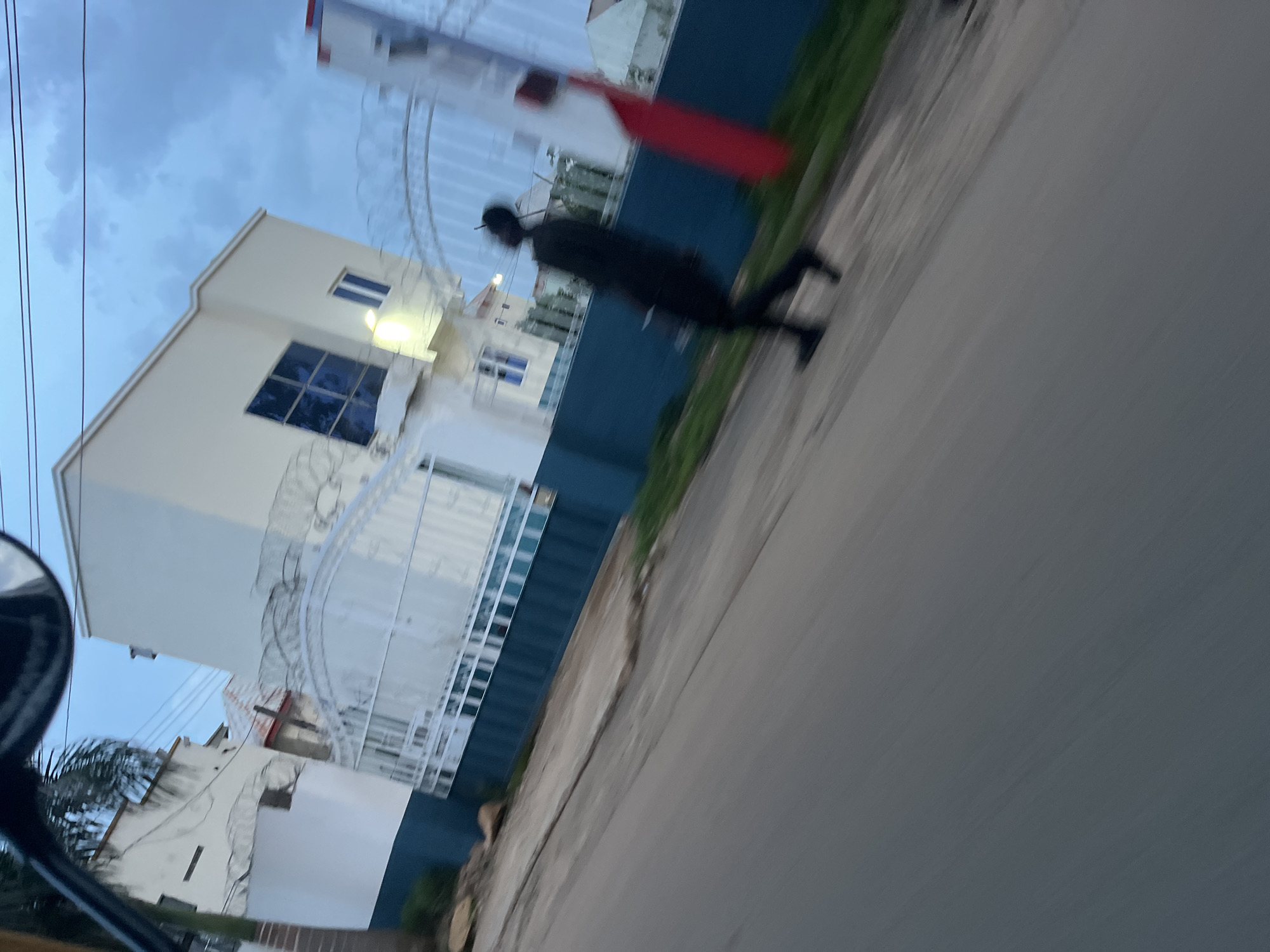 The image depicts a street scene in Bukuru, Nigeria, at dusk. A person dressed in black is walking on the right side of a paved road. In the background, a modern two-story building with a blue-tinted window is visible, bordered by a white gate and a dark blue fence. A tall, red and white structure is also present to the right of the building. The sky is overcast with scattered clouds. The perspective suggests the photo was taken from a moving vehicle, resulting in motion blur across the entire scene.