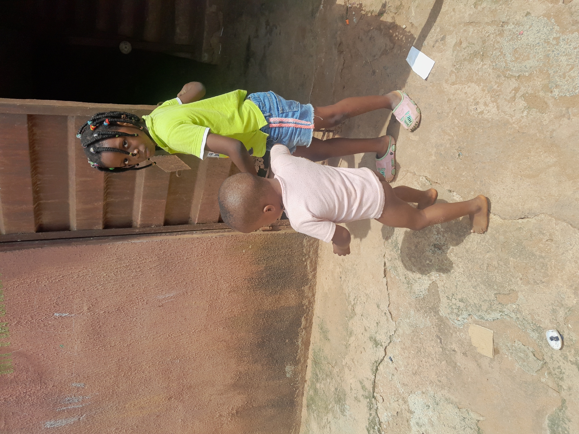 Two children, likely siblings, are positioned on a concrete surface outside a building in Nkpor, Nigeria. The older child, a girl, wears a neon green shirt and denim shorts with pink sandals. She has dark skin and braided hair with colorful beads. The younger child, possibly a toddler, is dressed in a light pink shirt and is barefoot. Both children are face-down on the ground. The girl looks at the camera. The location appears to be a simple outdoor setting, possibly a residential area. The wall of the building is visible. There are scattered objects on the ground, including a piece of paper and a small white object.