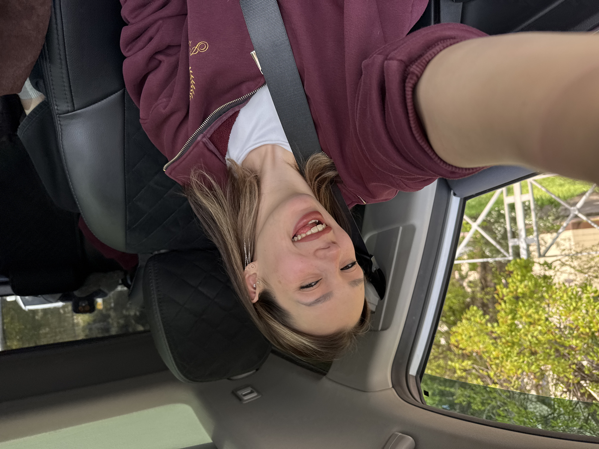 The image is a playful, upside-down selfie of a young person inside a car during the daytime. The individual, who appears to be female, has light brown hair with blonde highlights and is smiling broadly, sticking out their tongue. They are wearing a maroon or burgundy hooded sweatshirt over a white t-shirt, with a black seatbelt diagonally across their chest. The hoodie features a gold embroidered logo on the left chest area, which appears to be the stylized letters "ODDO" or "QUDO" surrounded by a wreath-like design. Several earrings are visible, including a longer, dangling chain earring on the left ear.
The car's interior features a black quilted leather seat behind the person, and the car's ceiling is visible above their head. Through the car window, lush green foliage and trees are visible, suggesting an outdoor setting, possibly suburban or rural. A light-colored structural element, possibly a fence or part of a building, can also be seen through the window. The bright natural light indicates it is daytime. The overall mood conveyed by the subject's expression is cheerful and lighthearted. The image is intentionally rotated 180 degrees.