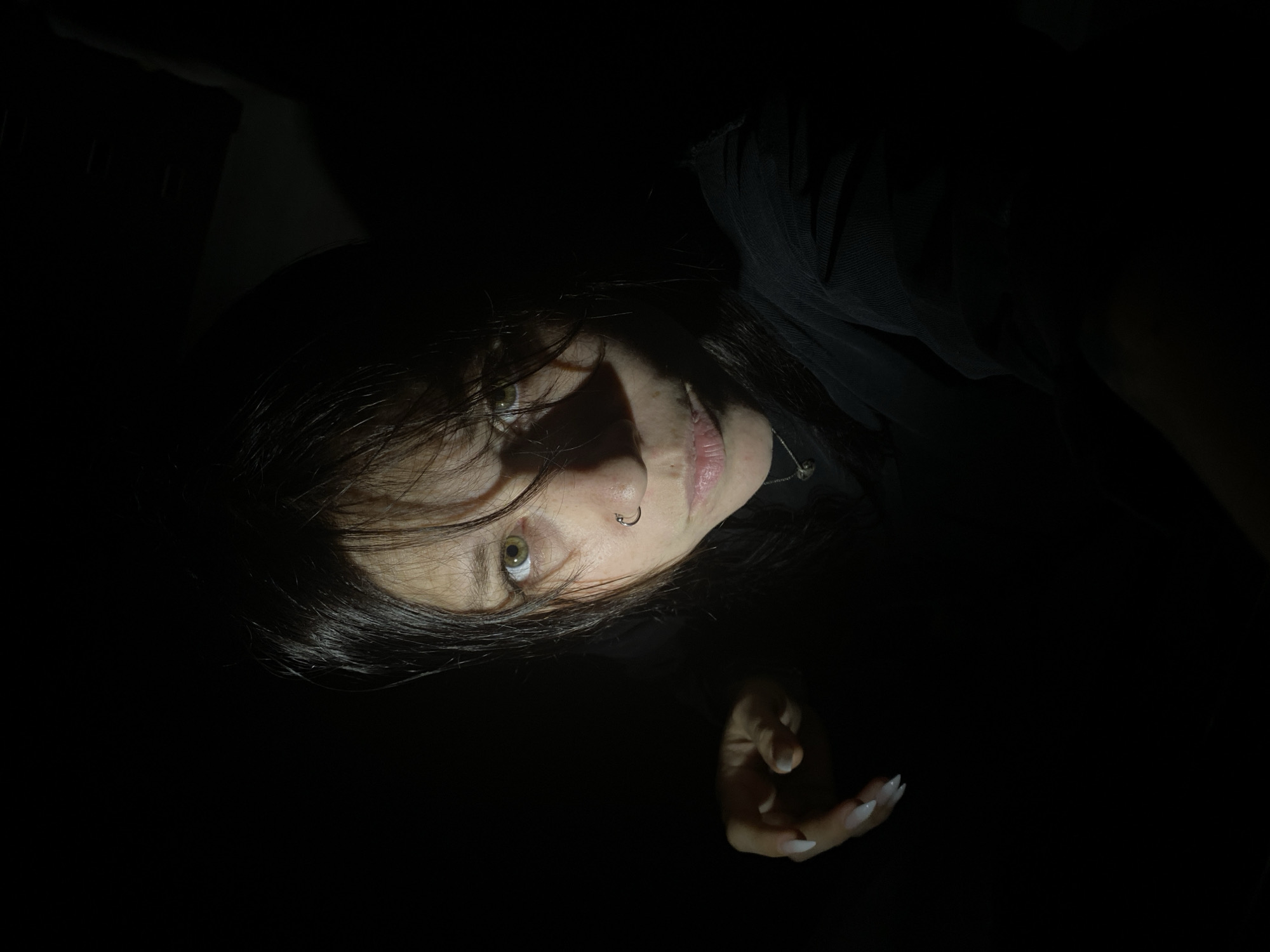 A person with dark hair and pale skin is lying down in a dark environment. The individual is wearing a black garment, and a piercing is visible on their nostril. One hand with long, pointed fingernails is partially visible in the lower right corner of the image. The location is not discernible, and no information indicates it is in Chaikovsky, Russia.
