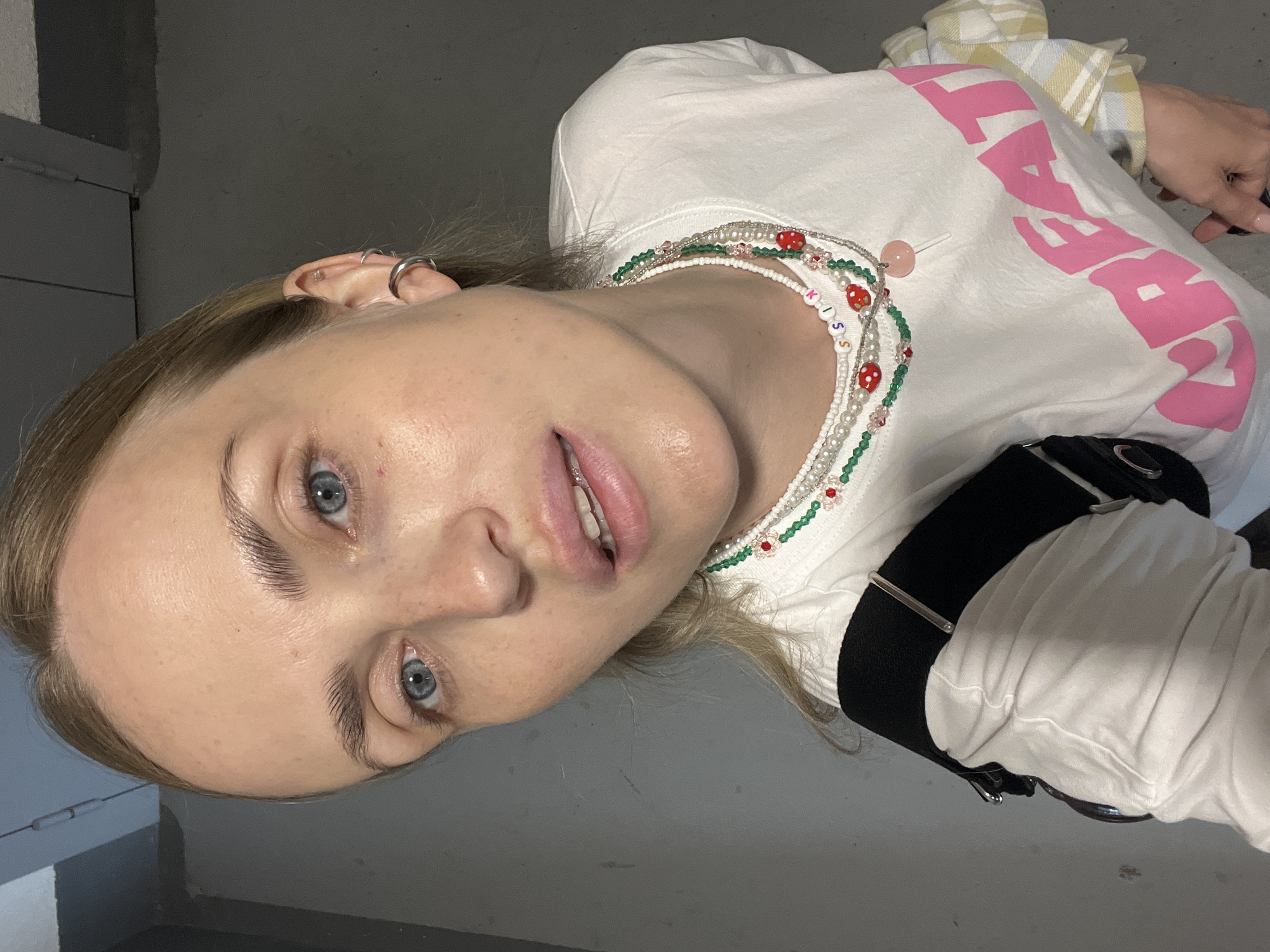 A selfie taken from a high angle shows a fair-skinned woman with blue eyes and blonde hair pulled back. She is wearing a white t-shirt with pink text on the front that appears to say "CREATE" and several layered necklaces, one of which has beads that spell out "SKIN" and strawberry charms. She has on a black strap across her chest. The background is a plain gray wall with some subtle texture. The lighting is dim and appears to be artificial. The woman has a nose ring and a silver hoop earring in her left ear. Her makeup is minimal, with a touch of lip gloss. She is indoors, possibly in a basement or industrial-looking space, given the concrete wall and muted lighting. No specific location in the city of Unknown, Russia can be identified.
