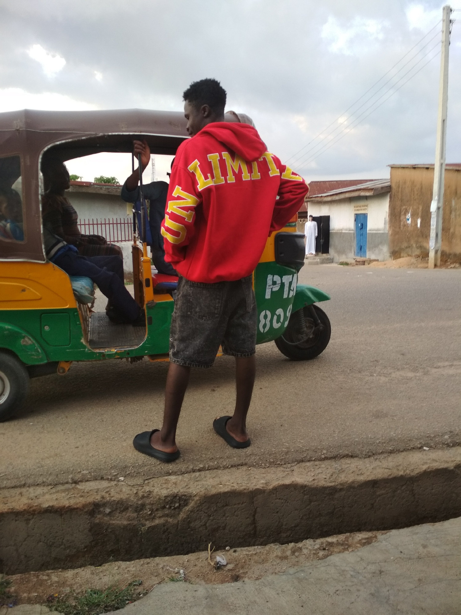 A medium outdoor shot depicts a person standing on a curb next to a three-wheeled vehicle, in an urban environment. The person is wearing a red hoodie with yellow lettering, gray shorts, and black sandals, facing away from the camera. Two individuals are seated inside the three-wheeled vehicle. A person in a white robe stands in the doorway of a building in the background. The sky is overcast. The vehicle has a green and yellow body with "PTA" and "809" painted on its side. A utility pole is visible to the right of the building.