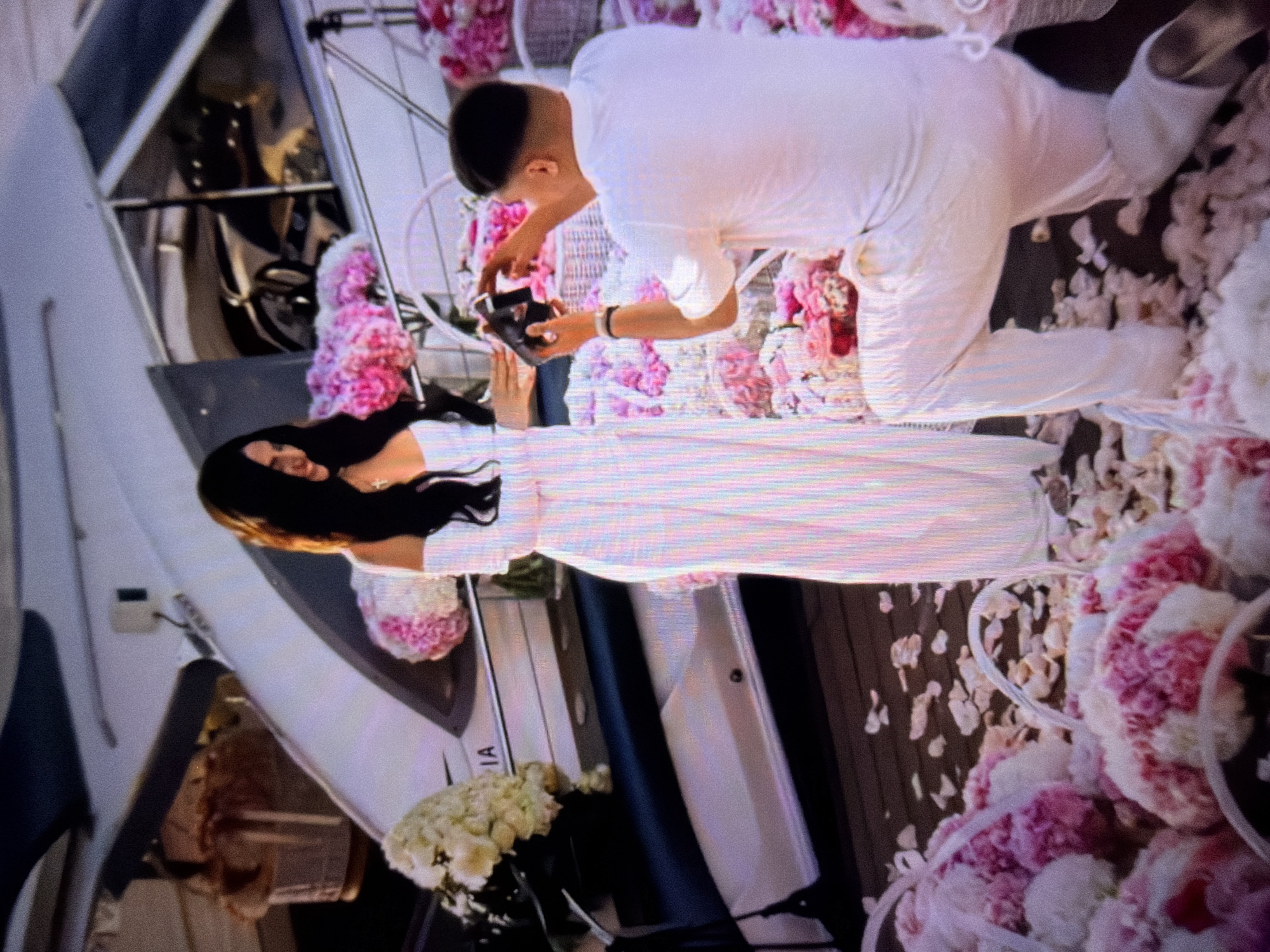 The media file shows a romantic scene likely set on a yacht or a similar vessel, decorated with an abundance of pink and white flowers. A man, dressed entirely in white, is on one knee, holding a small black object, possibly a ring box. He appears to be proposing to a woman who is kneeling or sitting before him. The woman, also wearing a white dress, has long dark hair and is looking down at the object in his hand. Her expression is difficult to discern clearly, but the setting suggests a moment of deep emotion and personal significance. The backdrop of the yacht is visible, with its sleek white exterior and some interior details. Scattered petals and floral arrangements create a luxurious and intimate atmosphere. The lighting suggests it could be daytime or early evening. The overall mood is one of love, celebration, and a significant life event, such as an engagement. There is visible text on the side of the yacht that reads "IA" followed by possibly more characters that are not fully legible.