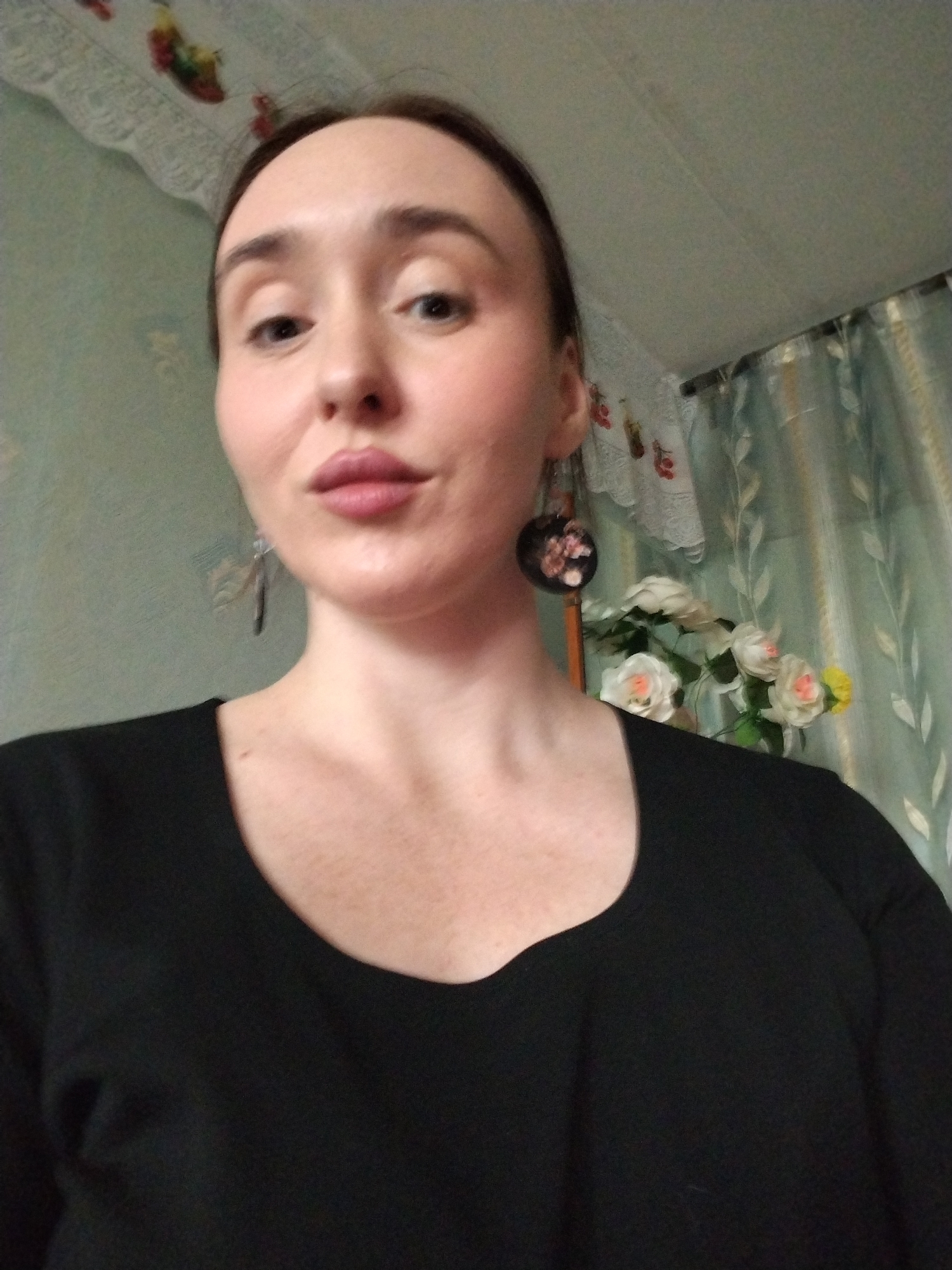 A Caucasian woman with light skin, reddish hair pulled back, and noticeable freckles is the central subject of this portrait-style image. She is wearing a black, scoop-necked shirt. She is wearing two earrings, one is a round, floral-patterned earring and the other a longer, more slender earring. Behind her, there is a backdrop featuring patterned wallpaper, lace trim, curtains, and artificial flowers. It appears to be an interior setting. The image was taken in Chaikovsky, Russia.