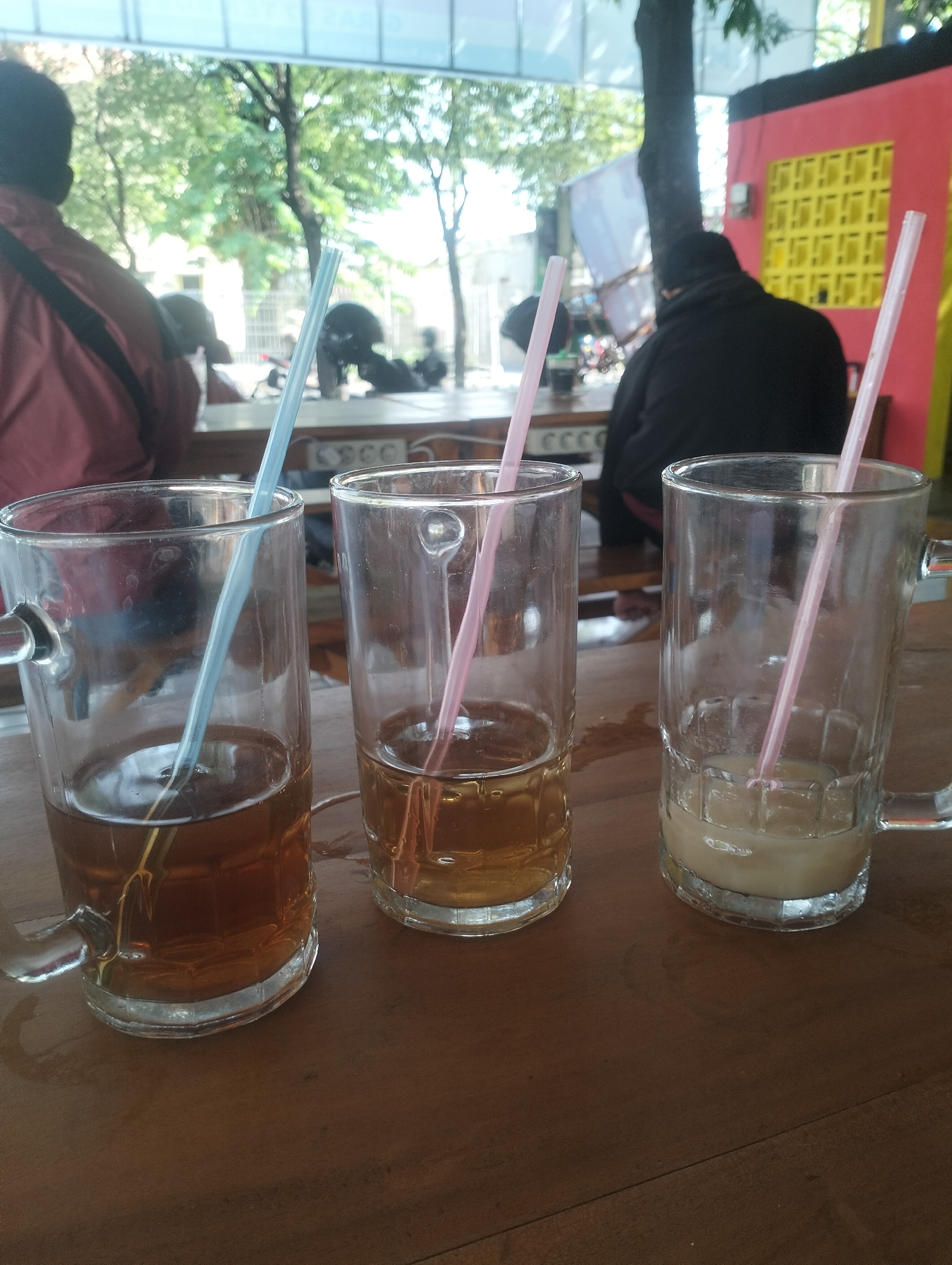 The image depicts three glasses with beverages and straws arranged on a wooden table. The glass on the left contains a dark amber liquid, possibly iced tea, with a light blue straw. The middle glass holds a lighter amber liquid, also with a pink straw. The glass on the right contains a pale, opaque liquid, possibly a creamy beverage, with a pink straw. In the background, blurred figures of people are visible, suggesting an outdoor seating area of a food establishment. The environment appears to be a public space with trees and a building in the distance. The overall scene suggests a casual setting, possibly a cafe or a roadside eatery in Surabaya, Indonesia, where people are enjoying drinks.