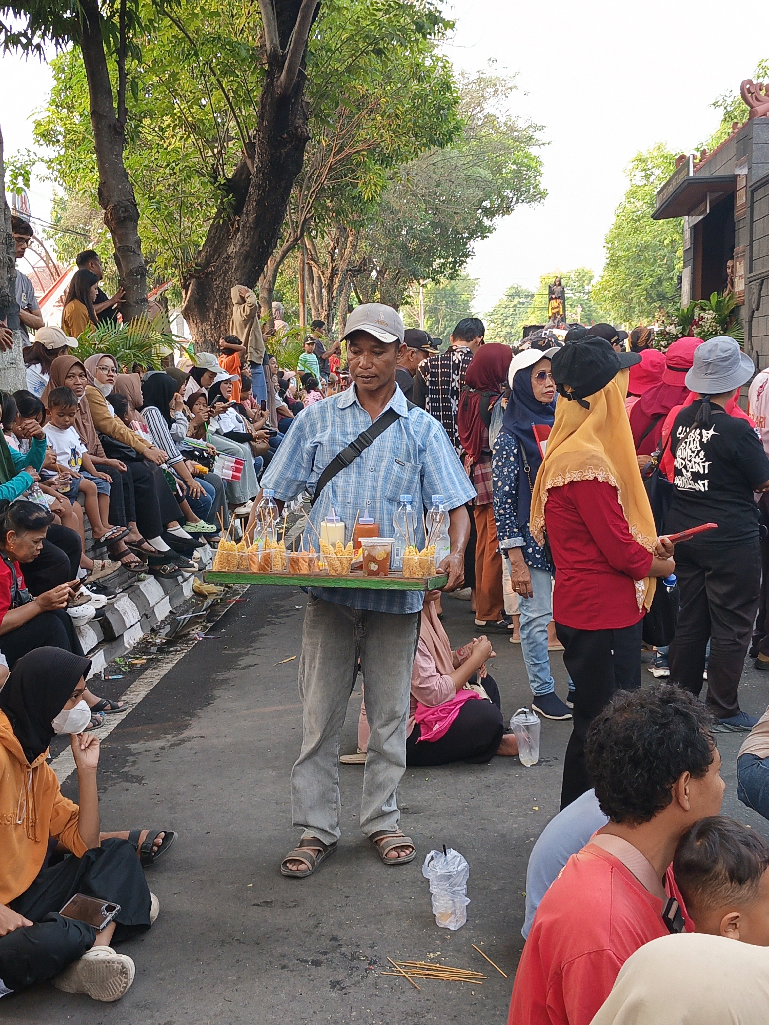 In the city of Gayamprit, Indonesia, a man in a blue checkered shirt and gray pants walks through a crowded street, carrying a tray of fried snacks. The tray contains several rows of food items, each skewered and presented upright. Accompanying the snacks are small containers of sauce and drinks. He walks on a paved road with a concrete barrier to his left, where a diverse group of people are seated, including women in hijabs and children. The background reveals more people, some standing and others sitting, suggesting a public gathering or event. Trees line the street, with lush green foliage indicating a warm climate. The sky is bright, suggesting it is daytime. The atmosphere appears to be lively, with many individuals present.