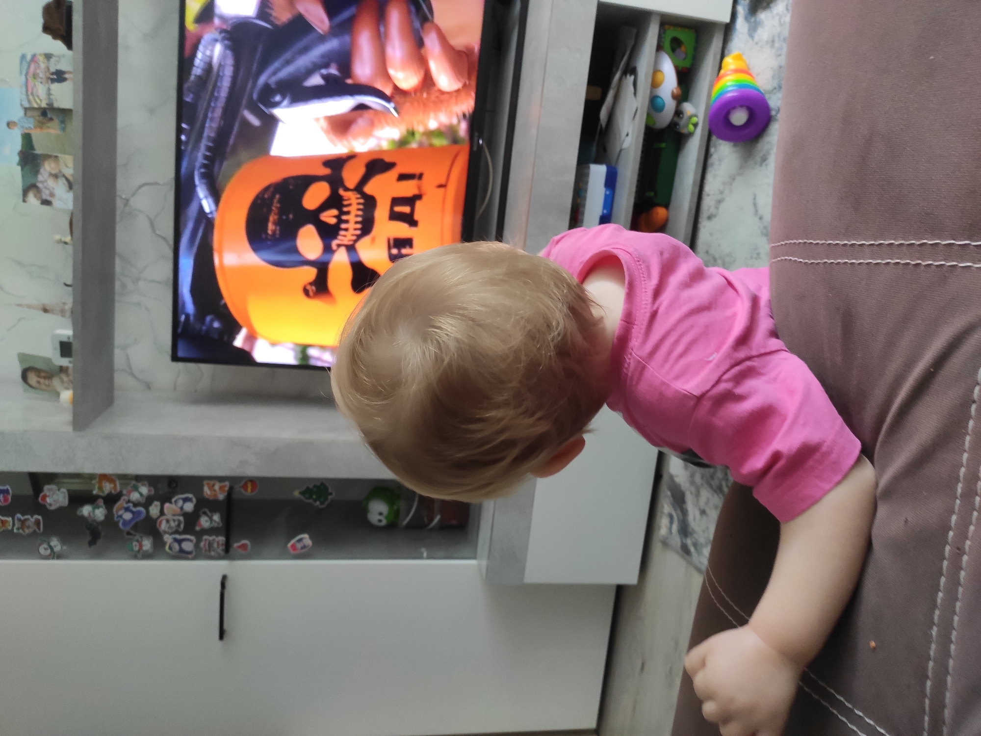 A young child with blonde hair, facing away from the camera, is seated on a brown couch, watching a television screen. The child wears a pink shirt. The television displays an orange bucket with a skull and crossbones symbol and Russian text. The environment appears to be a living room, with a white cabinet to the left featuring various stickers, and a media console below the television containing toys and other items. The floor is covered with a patterned rug. The overall scene depicts a domestic setting.