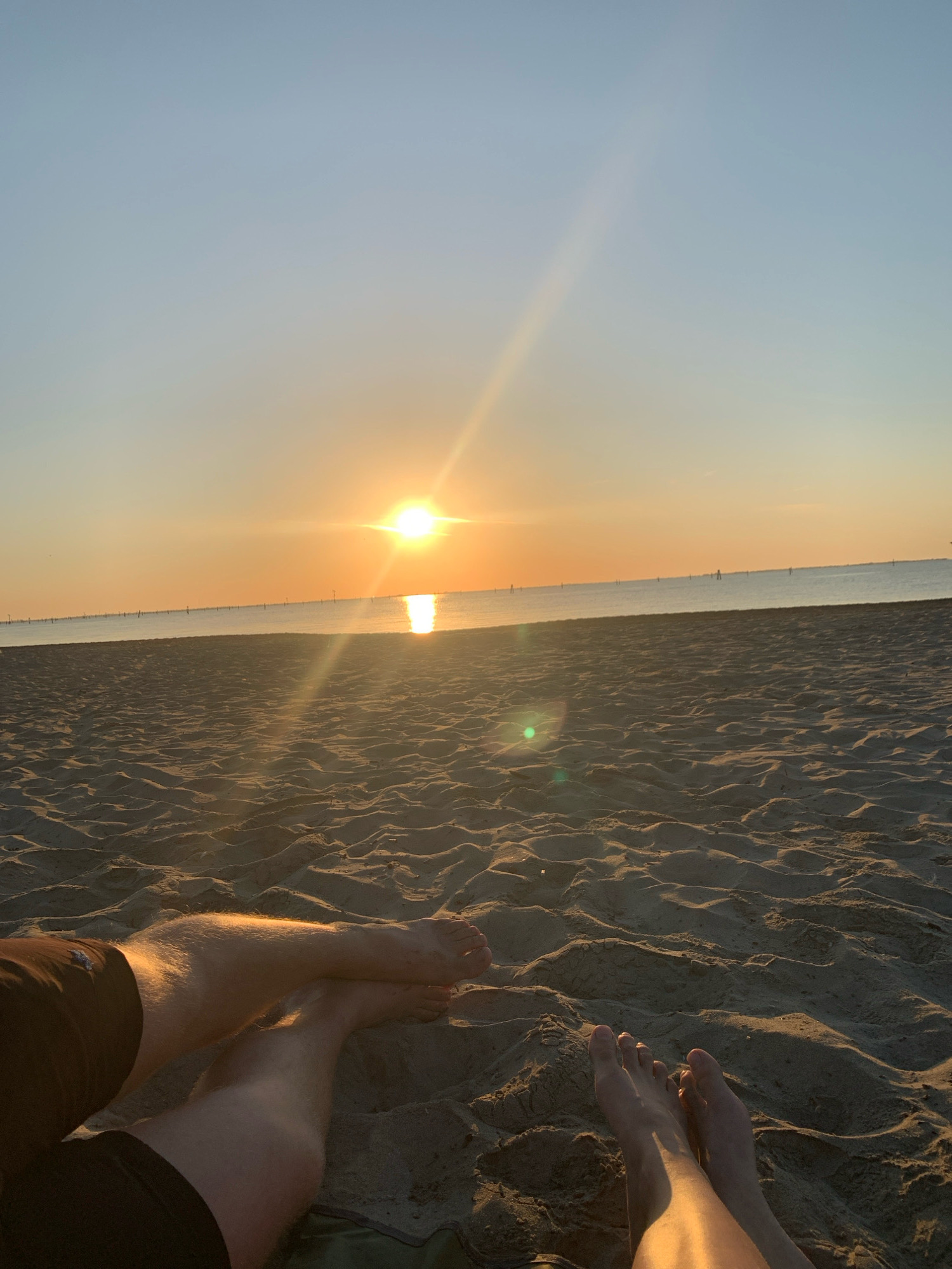 This image depicts a serene sunset scene on a sandy beach, presumably near the city of Comacchio, Italy. The sun, a bright orb, is low on the horizon, casting a warm, golden glow across the sky and the calm sea. The beach is covered in rippled sand, indicative of wind or water activity. In the foreground, the legs and feet of two individuals are visible, resting on the sand. One person's legs are crossed, with one foot resting on the other's thigh. The other person's feet are extended forward, toes slightly curled into the sand. The overall atmosphere is one of relaxation and tranquility, with the natural beauty of the sunset dominating the scene.
