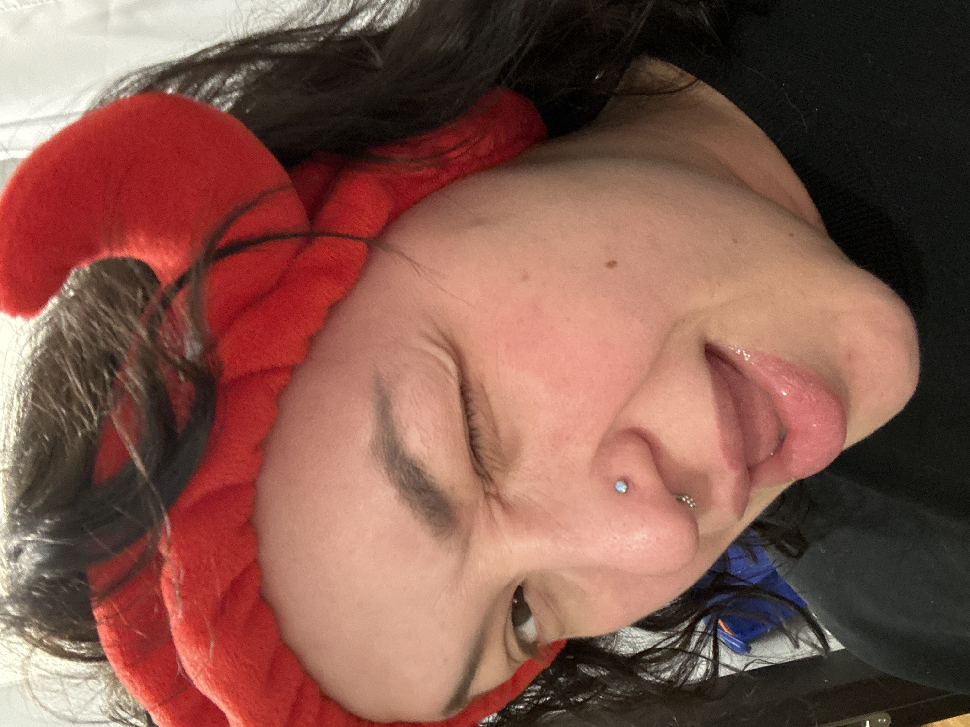 A close-up, angled shot depicts a person with dark hair wearing a red, plush headband with devil horns. The person's face is tilted towards the left. They have their tongue partially out. A silver nose piercing is visible on the left nostril. The environment appears to be indoors. There are no indications of the location being Chaikovsky, Russia.
