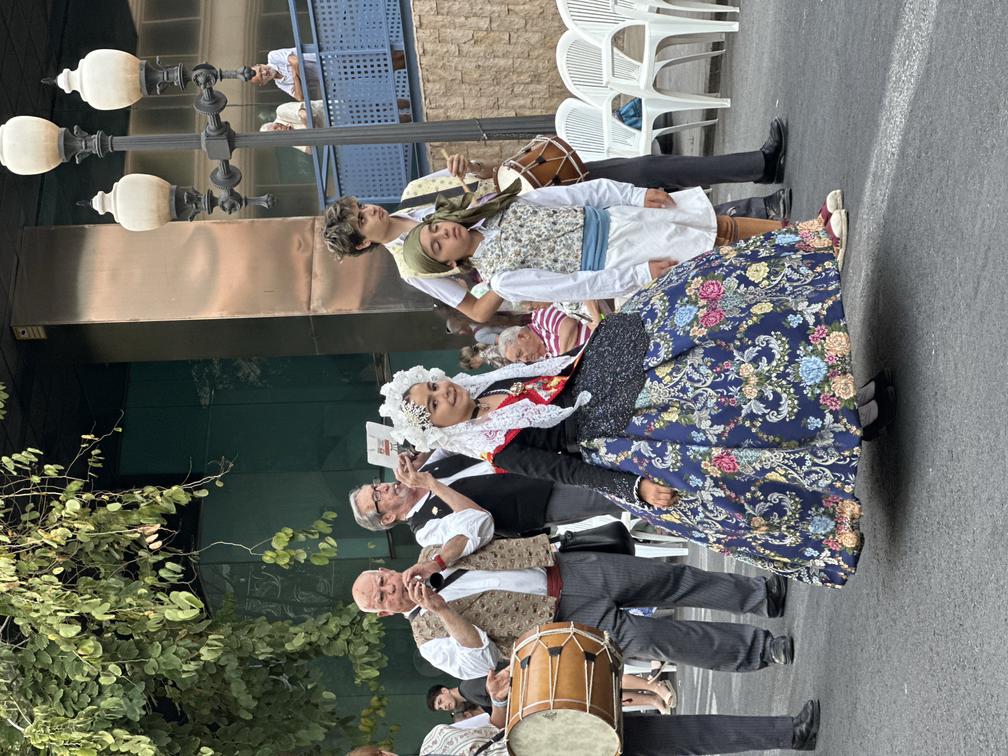 In a street scene, likely during a festival or parade, a group of people dressed in traditional attire are the central focus. In the foreground, a young girl wears an elaborate blue dress adorned with colorful floral patterns and a white lace headdress. To her left, an older man plays a wind instrument, while another man to her right also plays a similar instrument. A person on the far left is partially visible, holding a drum. Behind them, two young boys are also dressed in traditional clothing. The boy on the left is wearing a vest over a white shirt and pants, with a green bandana on his head, and is playing a drum. The boy on the right wears a similar outfit. In the background, white chairs are set up, suggesting an audience area, and people are standing on a raised walkway. Streetlights and trees are also visible, indicating an urban outdoor setting. The ground is paved, and a white line is painted on it.