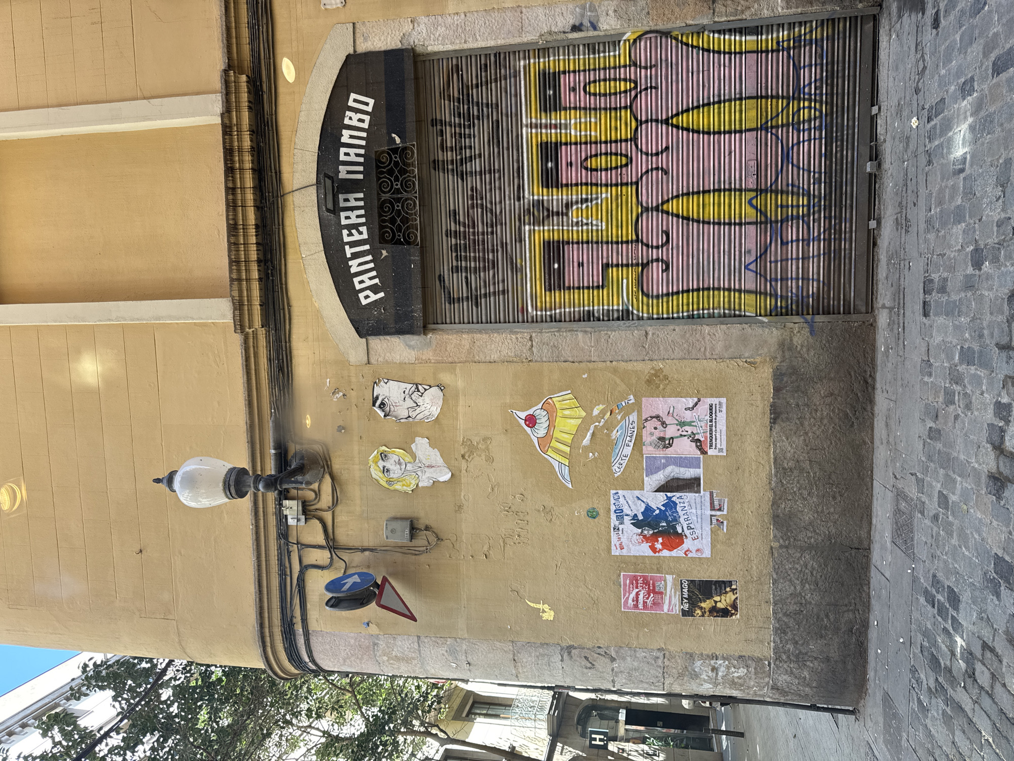 Exterior view of a building in Barcelona, Spain. The building facade is yellow with an arched doorway, which is sealed by a metal shutter covered in graffiti. The name "PANTERA MAMBO" is painted above the doorway. Various posters and stickers are affixed to the wall. A vintage street lamp is attached to the building, along with some signs. Cobblestone pavement is visible on the right side of the image.