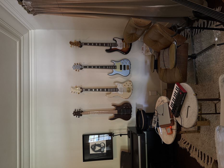 This image captures an interior room, likely a dedicated music space or living area, within the city of Los Angeles, United States of America. The exact street address or specific neighborhood is not discernible from the visual information provided.
The scene features a light-colored wall adorned with four electric bass guitars hung vertically. From left to right, these include a dark wood finished bass, a cream-colored bass with a pearloid pickguard and gold hardware, a light blue bass with a white pickguard and chrome hardware, and a sunburst finished bass with a white pickguard and chrome hardware.
To the left of the guitars, a black digital piano or synthesizer is partially visible. Above it, a framed black and white abstract artwork hangs, reflecting an outdoor view through a window, showing trees and natural light. A black stand, possibly for a microphone or sheet music, is positioned next to the piano.
In the central foreground, two nested, round tables with white marble tops and gold-colored metal bases are present. The upper table holds a silver laptop, an orange electronic device, a white power brick, and various cables. The lower table displays a black Akai MPK Mini MIDI keyboard controller with red accents and a small black hex wrench.
On the right side of the image, a brown leather Eames Lounge Chair is partially visible, featuring a black and white striped cushion. A tall, light-brown curtain hangs to the far right, suggesting a window. A black guitar stand is situated in the bottom right corner, holding the neck portion of a dark string instrument. A white electrical outlet is visible on the wall below the mounted guitars. No individuals are present in the scene. The arrangement of instruments and electronic equipment indicates an environment set up for musical production or appreciation.