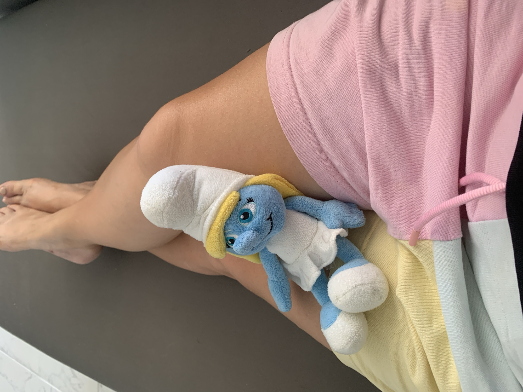 A Smurf plush toy rests on a person's crossed legs, which are clad in a pair of pastel-colored shorts. The person's bare feet are visible at the top of the frame. The background consists of a dark gray surface, possibly a lounge chair or sofa. The image was captured in Antalya, Turkey.