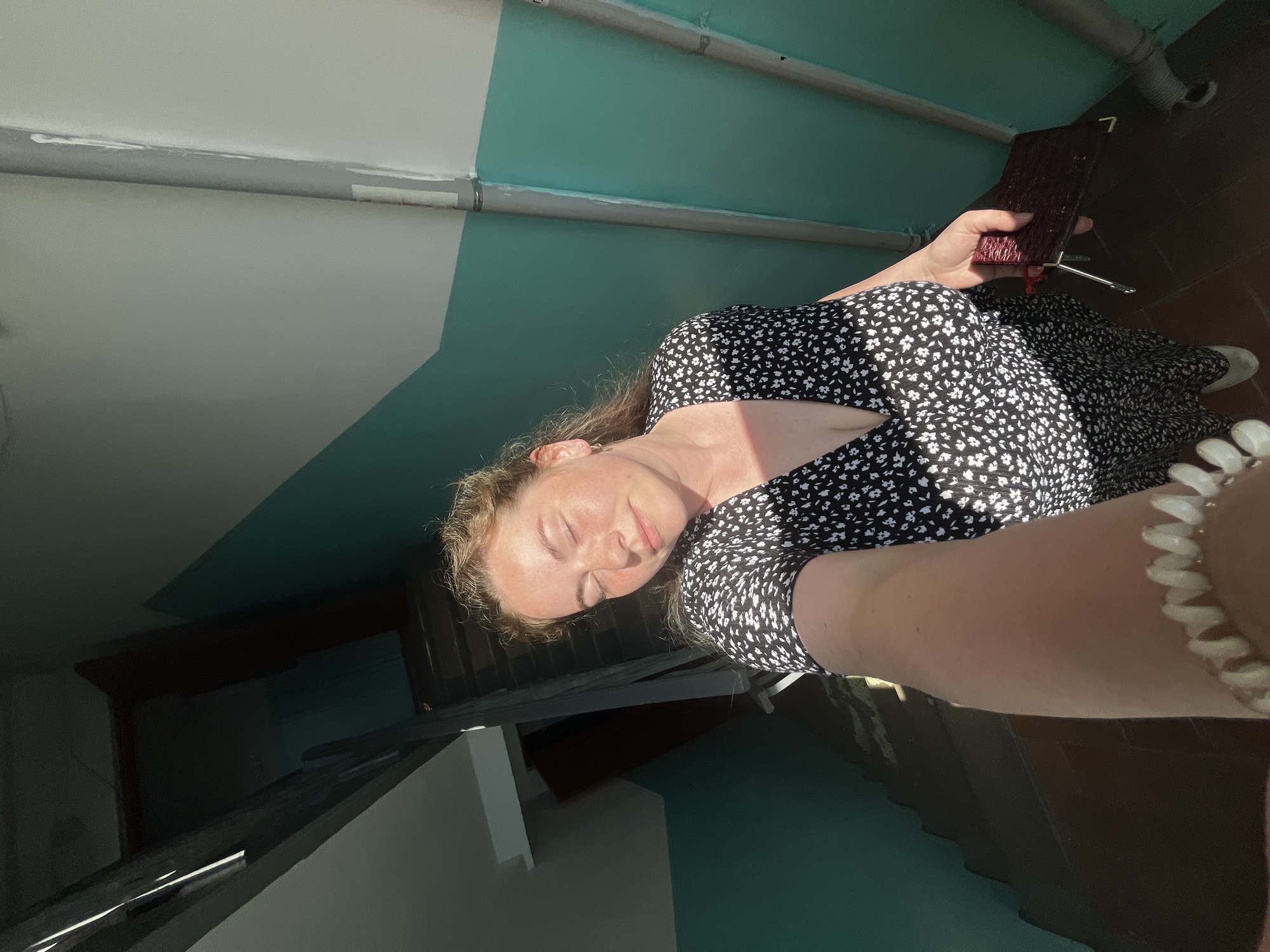 A woman with fair skin and blonde hair, wearing a black and white floral dress and a beaded bracelet, is lying down and holding a small red book or wallet. She is outdoors on a landing with a teal and gray wall. The location is indicated to be in the city of Chaikovsky, Russia. The floor is brown, and sunlight is illuminating the scene. The woman's eyes are closed as if she is resting.