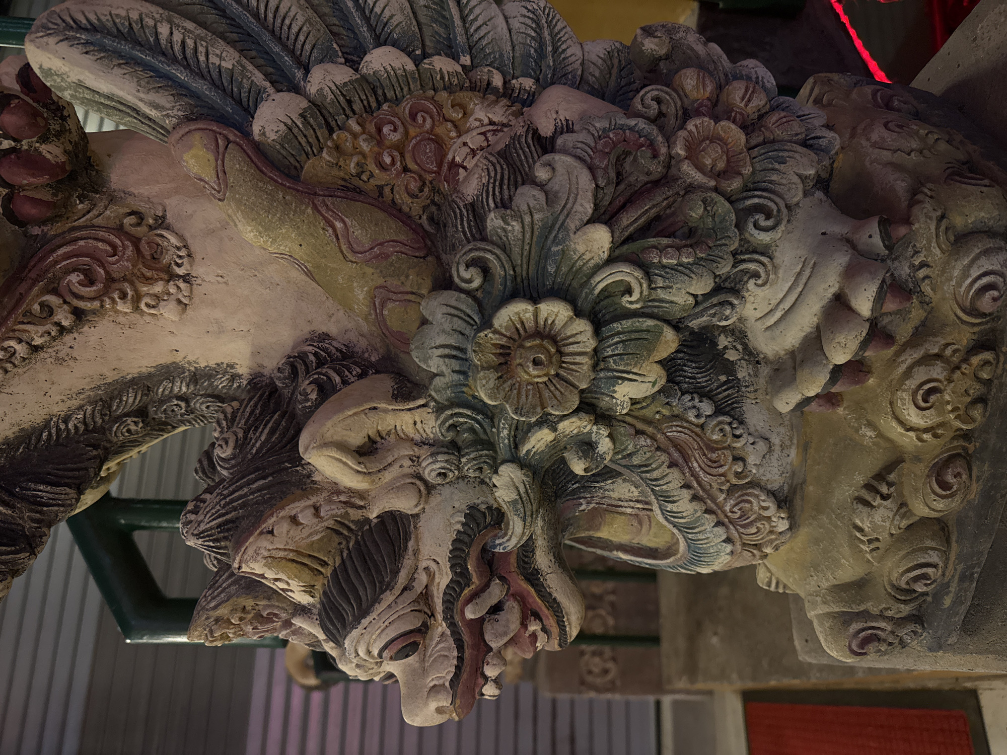 A close-up, eye-level shot shows a detailed, weathered stone statue of a mythical creature, likely a garuda or dragon, in Kuta, Indonesia. The statue is predominantly beige with accents of red, blue, and green paint, showing signs of age and wear. The creature has an open mouth with sharp teeth, a scaly texture, and a prominent winged design. It stands on a decorative base. In the background, a green metal railing and a corrugated metal wall are visible, suggesting an outdoor or semi-outdoor location. A red object, possibly a mat, is partially visible in the lower left corner.