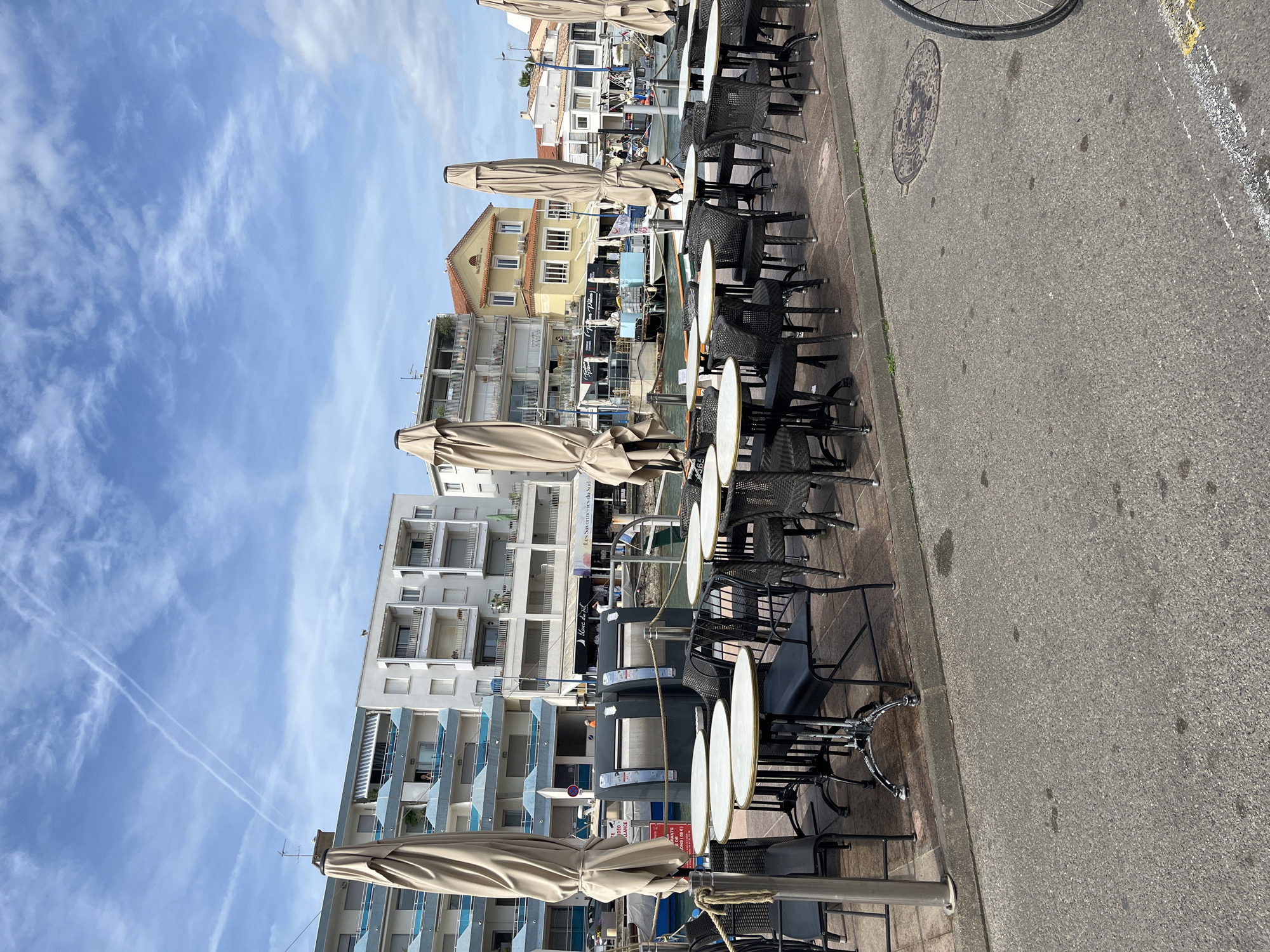 This bright daylight scene captures a tranquil waterside promenade in Palavas-les-Flots, France. A row of neatly arranged outdoor cafe seating lines the paved sidewalk on the right, featuring numerous black wicker-style chairs and small, round white tables, many shaded by large, beige parasols. No people are actively visible at the cafe tables, suggesting a quiet moment.
Beyond the cafe area, on the left, a narrow body of water, likely a marina or canal, is visible, with several boats docked. The far side of the water is flanked by a mix of buildings: modern multi-story structures with balconies, and older, traditional-style buildings with light yellow facades and red-tiled roofs.
The upper part of the image shows a clear blue sky with scattered wispy white clouds and prominent airplane contrails. On the asphalt road to the far right, a manhole cover with an intricate design is visible, along with the partial wheel of a bicycle. The bright sunlight casts soft shadows, indicating it is daytime.
Visible text details include "Les Savoureuses du Sud" and "Fusions" on building signs, and a red sign with French warnings regarding alcohol: "TOUTE ALCOOLISE OU..." (likely "any intoxicated person or...") and "VENTE AUX MINEURS DE..." (likely "sale to minors under..."). The scene evokes a peaceful, sunny day in a charming coastal town.