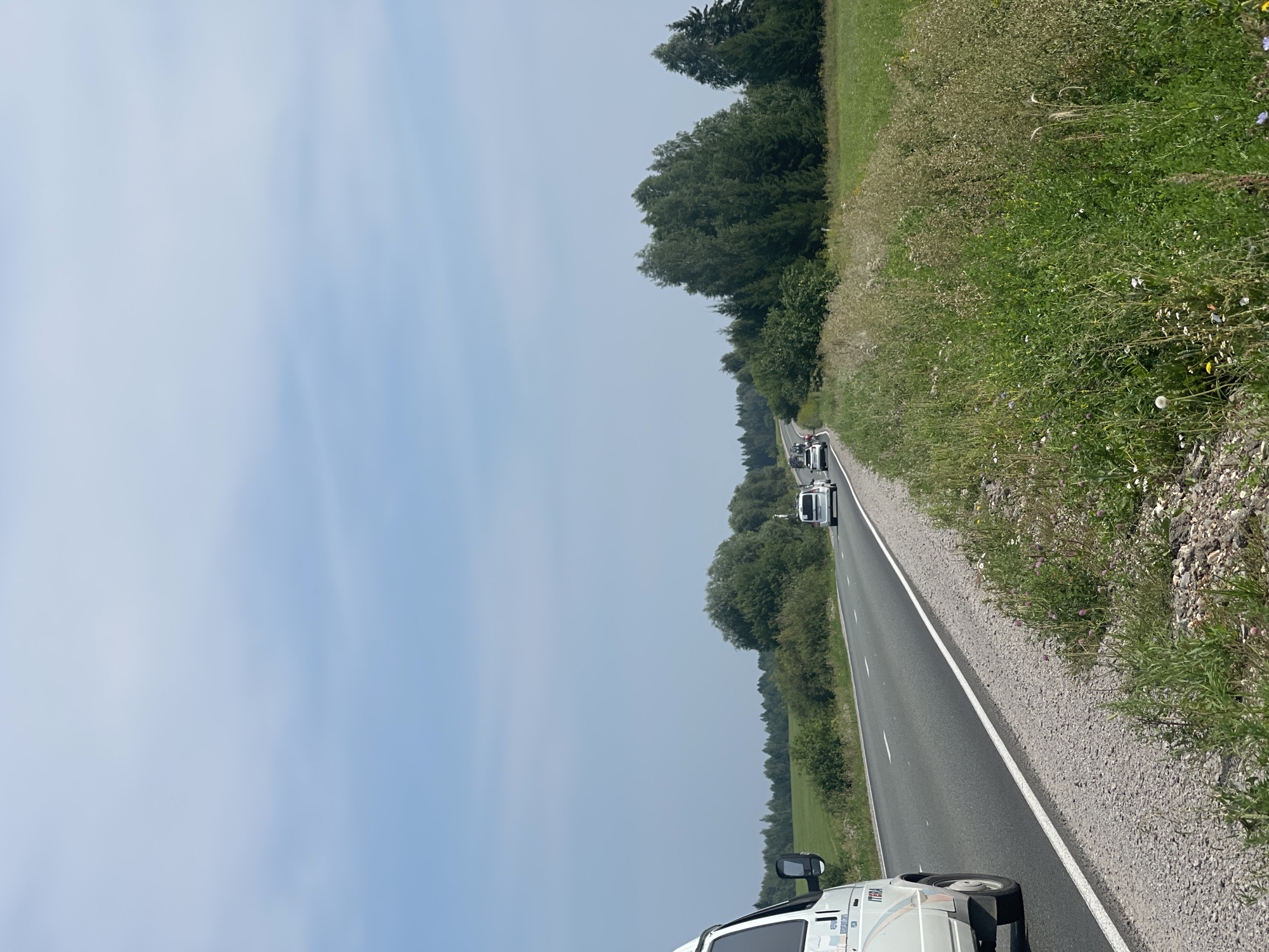 The image shows a sunny day in Perevoznoe, Russia, with a clear blue sky and scattered clouds. A white van is parked on the left side of a paved road, with its rear facing the viewer. The van has "ITERA" written on its side. Several other vehicles, including a white minivan and a few cars, are driving on the road, heading away from the viewer and disappearing around a bend.
The road is bordered by a grassy shoulder with wildflowers and tall grass. On the right side of the road, a dense forest of green trees stretches into the distance. The landscape appears rural and natural, with no buildings or signs of urban development visible. The overall scene suggests a quiet, scenic drive through the countryside.