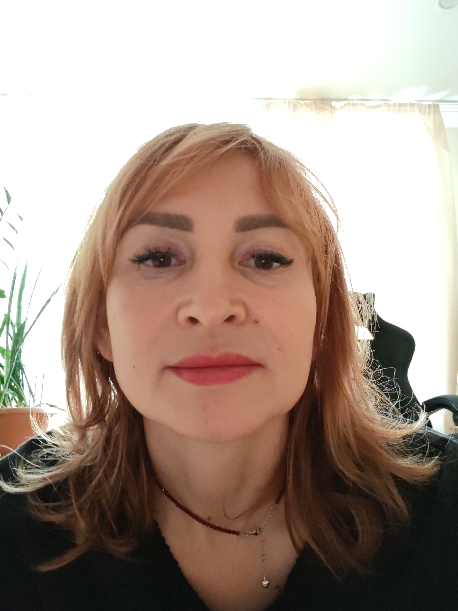 A fair-skinned woman with light brown hair and a red necklace is centered in a close-up shot. She is wearing dark clothing and light makeup. A plant is partially visible on the left, and the back of an office chair is on the right. A bright window is behind her, suggesting an indoor setting. There is no visible information to ascertain that this photograph was taken in the city of Chaikovsky, Russia.