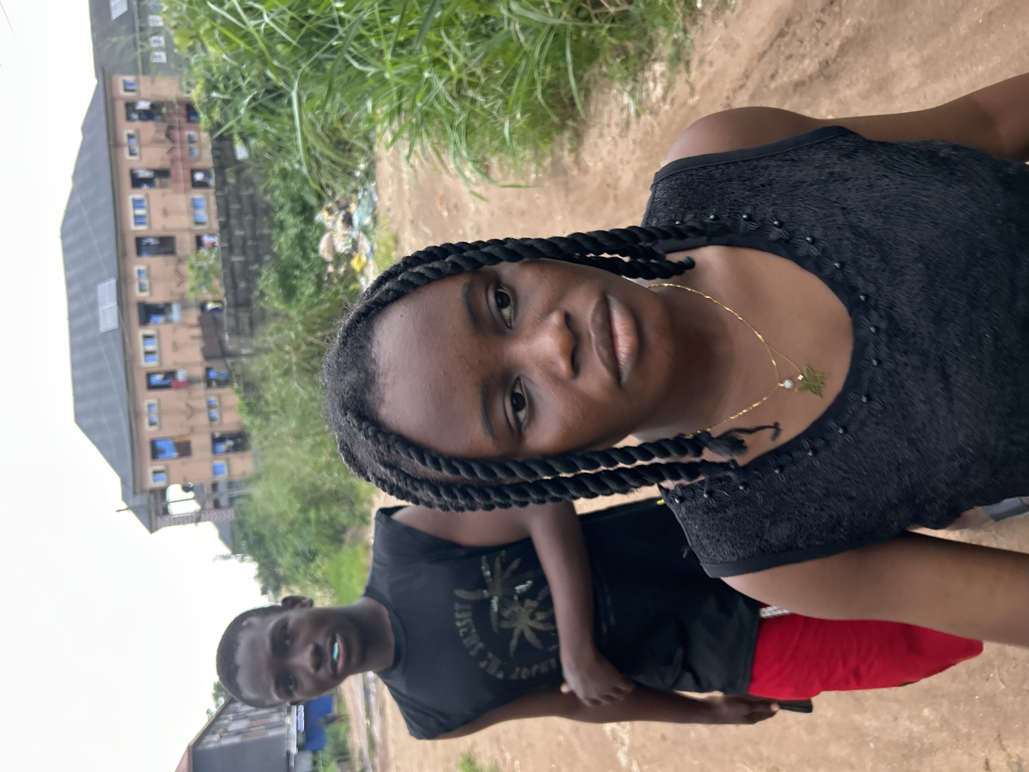 The image depicts a vertical medium shot of two young Black individuals outdoors in Ihiagwa, Nigeria. A young woman with dark, braided hair is in the foreground, facing the camera with a neutral expression. She is wearing a black, sleeveless top adorned with decorative beadwork and a delicate gold necklace with a star pendant.
Behind her, a young man stands slightly to the left, also facing the camera. He is wearing a black t-shirt with a graphic design and red shorts. His mouth is slightly open, revealing a light blue object, possibly gum or candy.
The setting appears to be a somewhat informal outdoor area, with a dirt path and overgrown green vegetation in the background. To the far left, a multi-story building with visible windows and balconies can be seen, suggesting an urban or semi-urban environment. There is also a pile of what appears to be trash or debris near the building.
The lighting suggests it is daytime, with the sun casting a soft illumination. No specific weather conditions are evident, though the vegetation implies it might be a humid or rainy season. The overall mood is casual and candid. There is no visible text in the image.