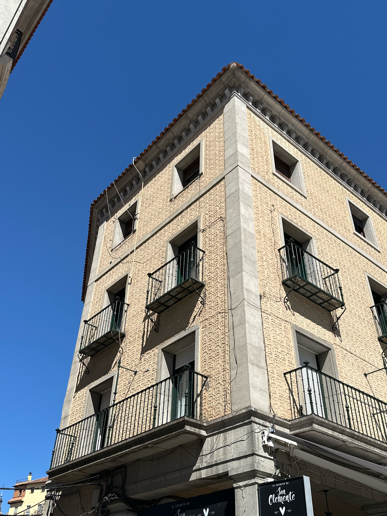 The image shows a corner view of a building with a yellow facade adorned with an intricate embossed pattern. The building has several windows, some with wrought-iron balconies, and a tiled roof. The sky is a clear blue. The architecture suggests it could be in an older European city, possibly Segovia, Spain, given its historical building styles often feature such decorative facades and balconies. The building appears to be multi-story. There are no people visible in the immediate frame. The lower portion of the building shows a sign for "San Clemente" and possibly a cafe or restaurant. Electrical wires are visible running along the exterior of the building.