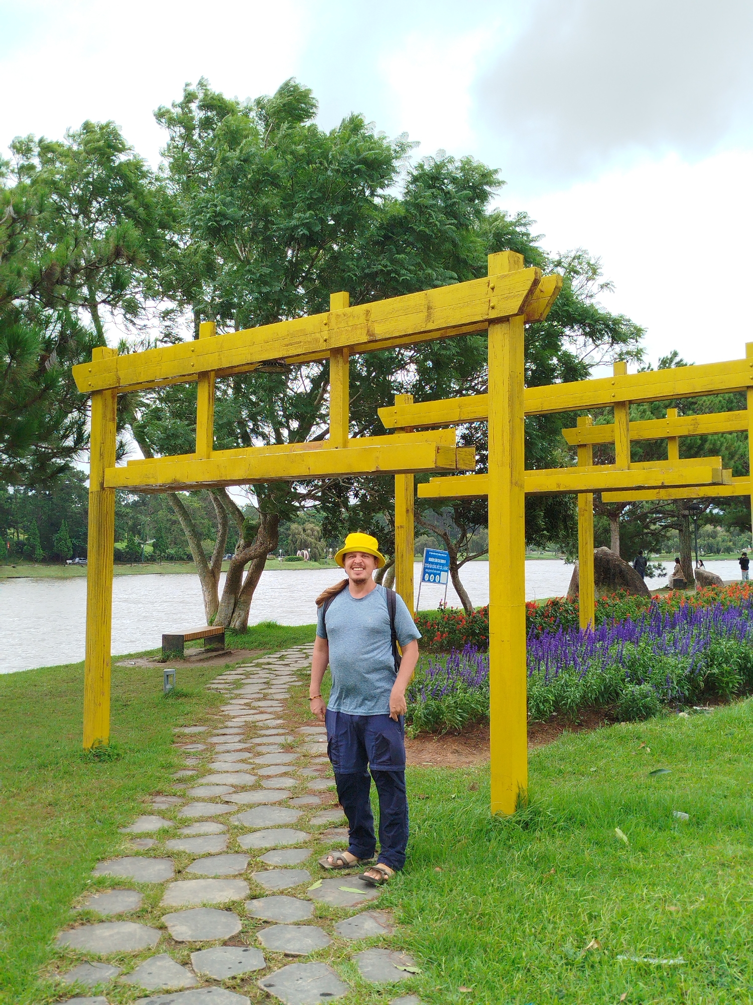 A man wearing a yellow bucket hat and a blue t-shirt stands on a stone path in a park. He is smiling and looking at the camera. Behind him, there is a yellow wooden structure resembling a torii gate, a body of water, lush green trees, and a flower bed with purple flowers. The scene appears to be in Da Lat, Vietnam, likely at the Tuyen Lam Lake area, known for its scenic beauty and recreational activities. The man is wearing dark blue cargo pants and sandals. A backpack is visible on his shoulders. The sky is overcast.