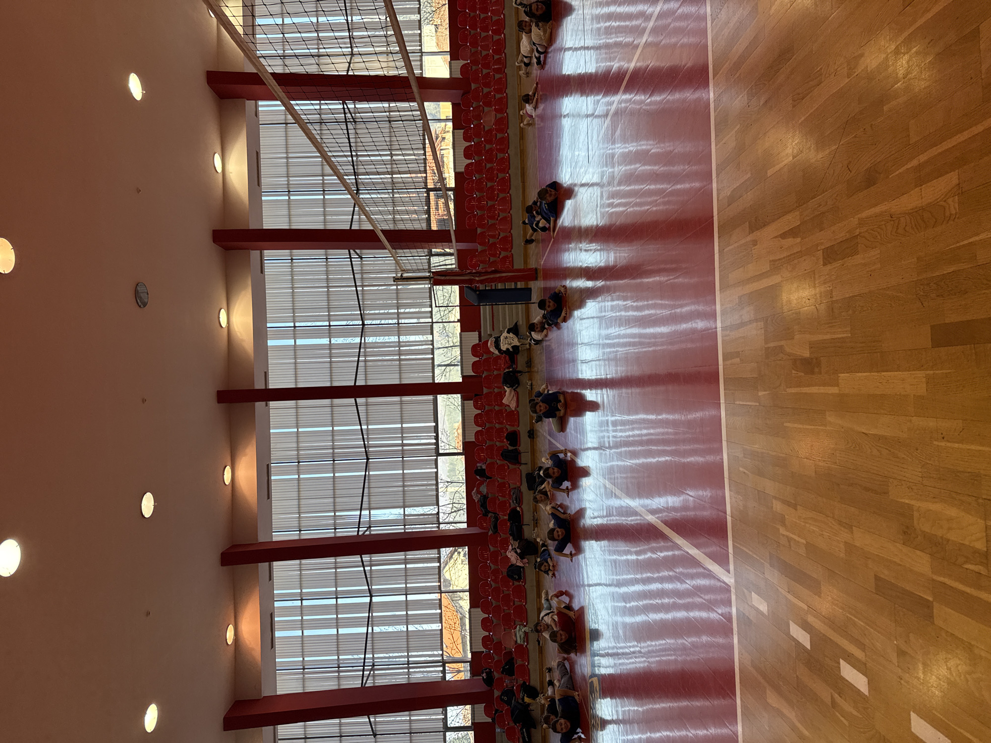 The image shows an indoor gymnasium in Etropole, Bulgaria. The floor is partially red and partially varnished wood, marked with white lines. Rows of red seating line one side of the gym. A large window runs along the top of the seating, providing natural light. A volleyball net is present in the center of the room. A group of people are sitting along the edge of the court, some facing the seating area, others facing the court. The walls are light-colored with evenly spaced circular lights.