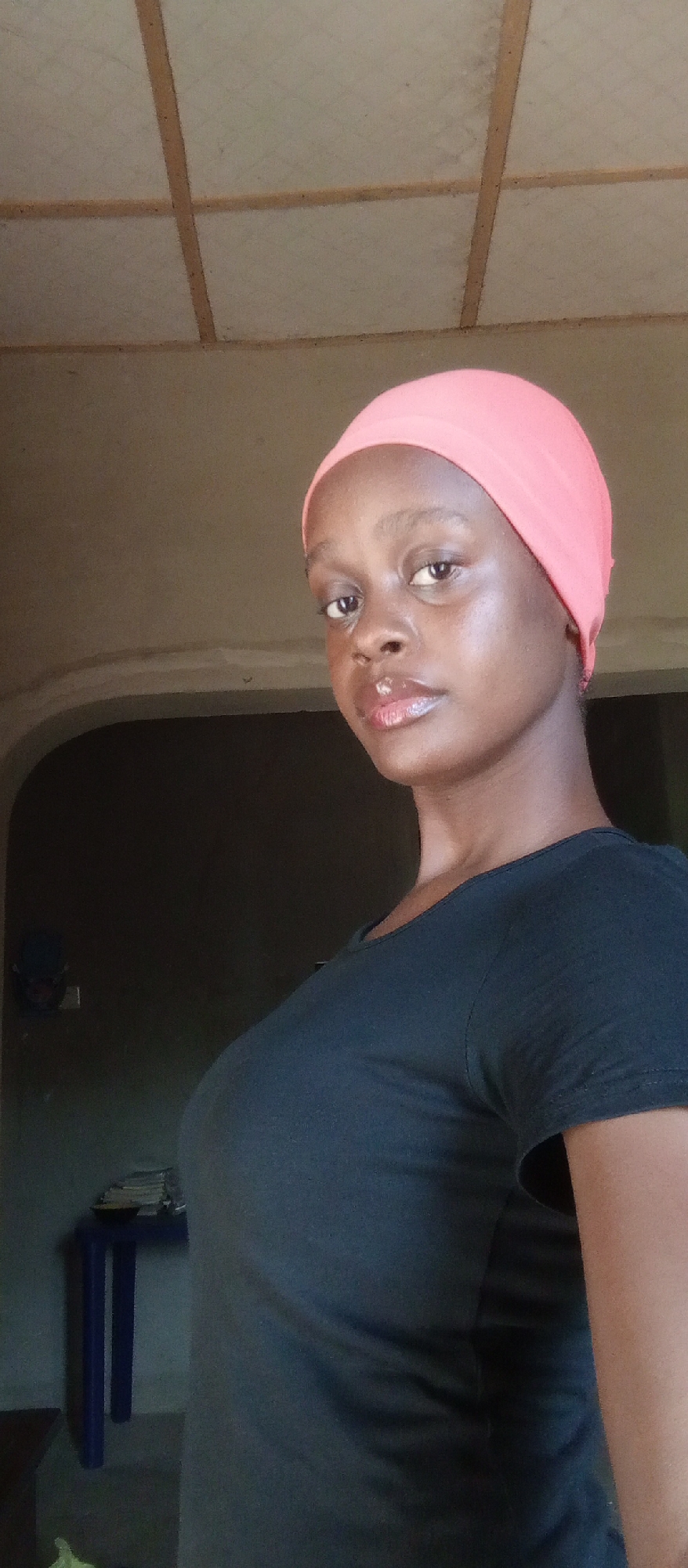 A young woman with dark skin and glossy lips wears a pink head wrap and a dark blue or black t-shirt. She looks towards the camera with a serious expression. The background includes a room interior with a plain, light-colored ceiling divided into squares. Behind the woman, a dark doorway or archway is visible, and a small blue table with items on top sits near the doorway. The location is likely in a home or residential building in Akwanga, Nigeria, based on the general appearance and style of the interior.