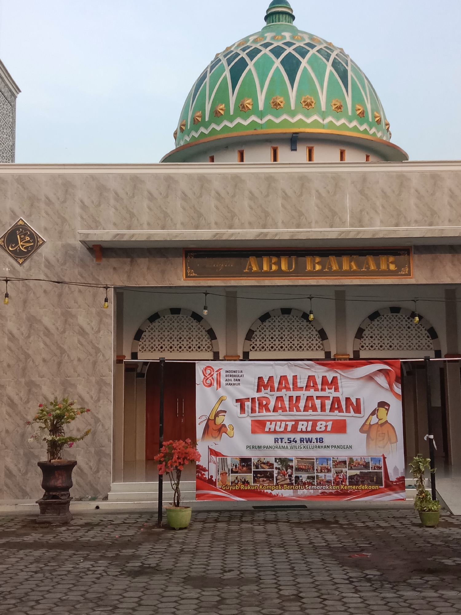 The image shows the facade of a building identified by the sign "ABU BAKAR" above the entrance. The building has a large, ornate green dome with gold accents and a patterned design. Below the dome, the building is clad in marble-like tiles. A red and white banner is prominently displayed in front of the entrance, featuring text and images related to an event called "MALAM TIRAKATAN HUT RI 81". The banner indicates the location as "RT.54 RW.18 Perum Panorama Wilis Kelurahan Pandean". The ground in front of the building is paved with interlocking gray bricks, and several potted plants are visible. The scene is captured during daylight, with a slightly overcast sky.