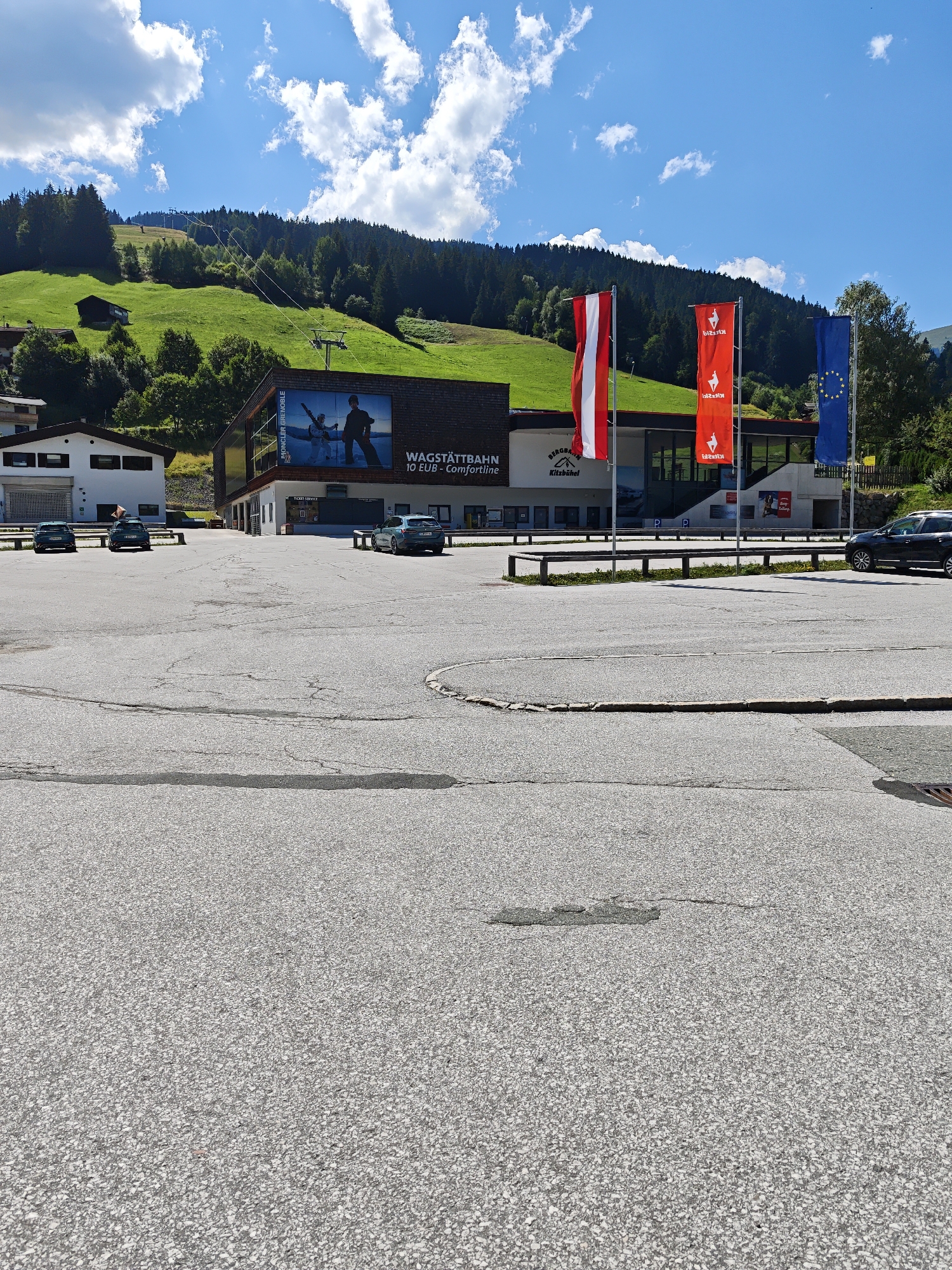 A sun-drenched scene unfolds in Filzen, Austria, featuring a modern building facade adorned with flags and a large mural. The building, likely a transportation hub or tourist facility, displays prominent signage indicating "WAGSTATTBAN" and "10 EUR - Comfortline." Three vertical flags, representing Austria, a red flag with a white goat emblem, and the European Union, flutter in the breeze. Behind the building, a lush green hillside rises, dotted with trees and a ski lift, hinting at winter sports activities. Several cars are parked in the foreground on an asphalt lot, their presence suggesting a public access area. The sky is a brilliant blue, punctuated by fluffy white clouds.