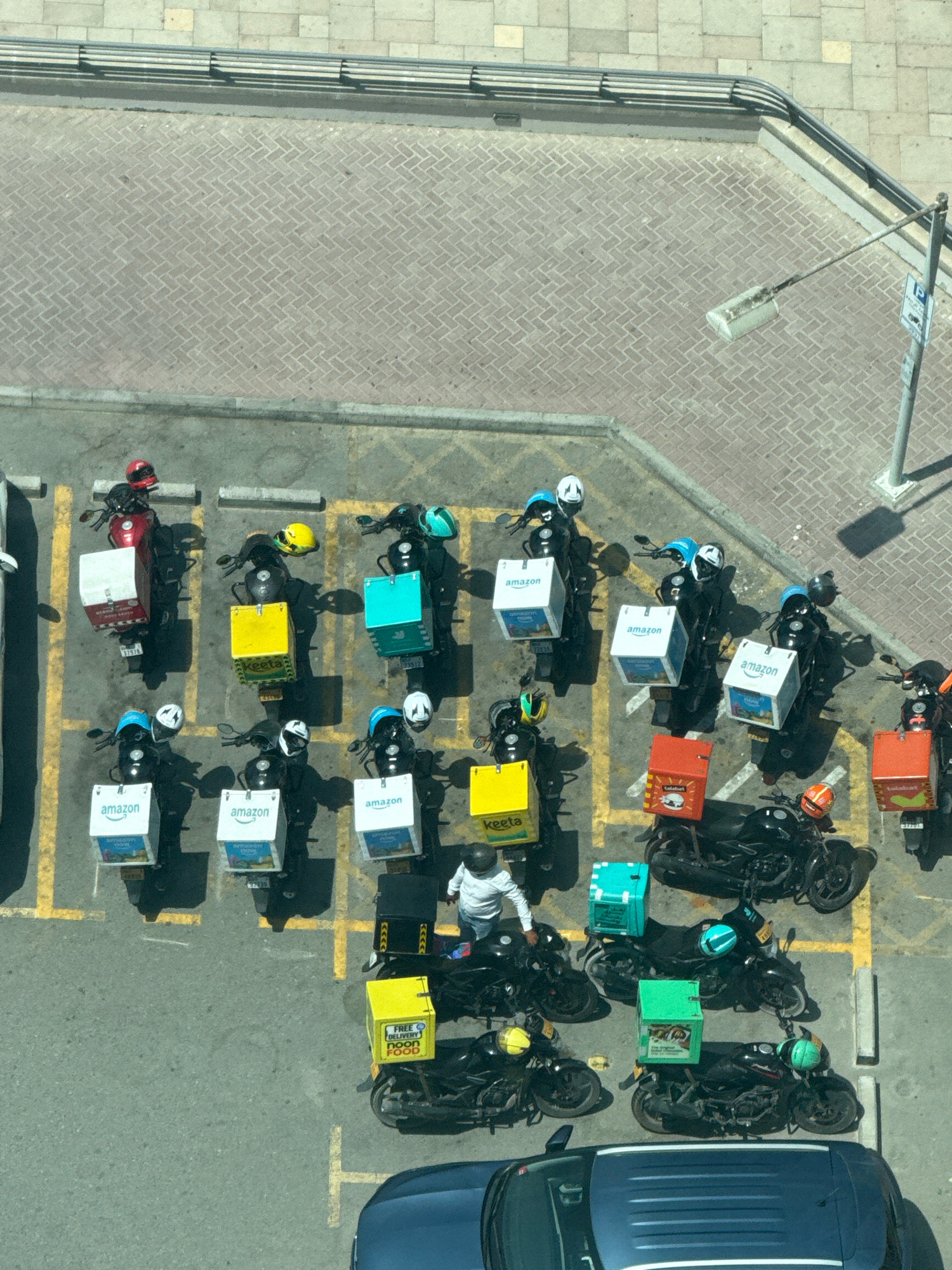 An aerial view shows a parking lot in Dubai, United Arab Emirates, filled with delivery motorcycles. The motorcycles are arranged in rows, each equipped with a storage box on the back. The boxes vary in color and branding, with some displaying the "amazon" logo, others labeled "keeta," and some with unidentifiable red or green branding. Several motorcycles have helmets placed on them, and a single individual wearing a white long-sleeved shirt is standing next to one of the motorcycles. The parking lot is paved with interlocking bricks and delineated by yellow lines. In the background, a street with a sidewalk and a metal railing is visible, with a lamppost and a parking sign. A portion of a dark blue vehicle is in the foreground.