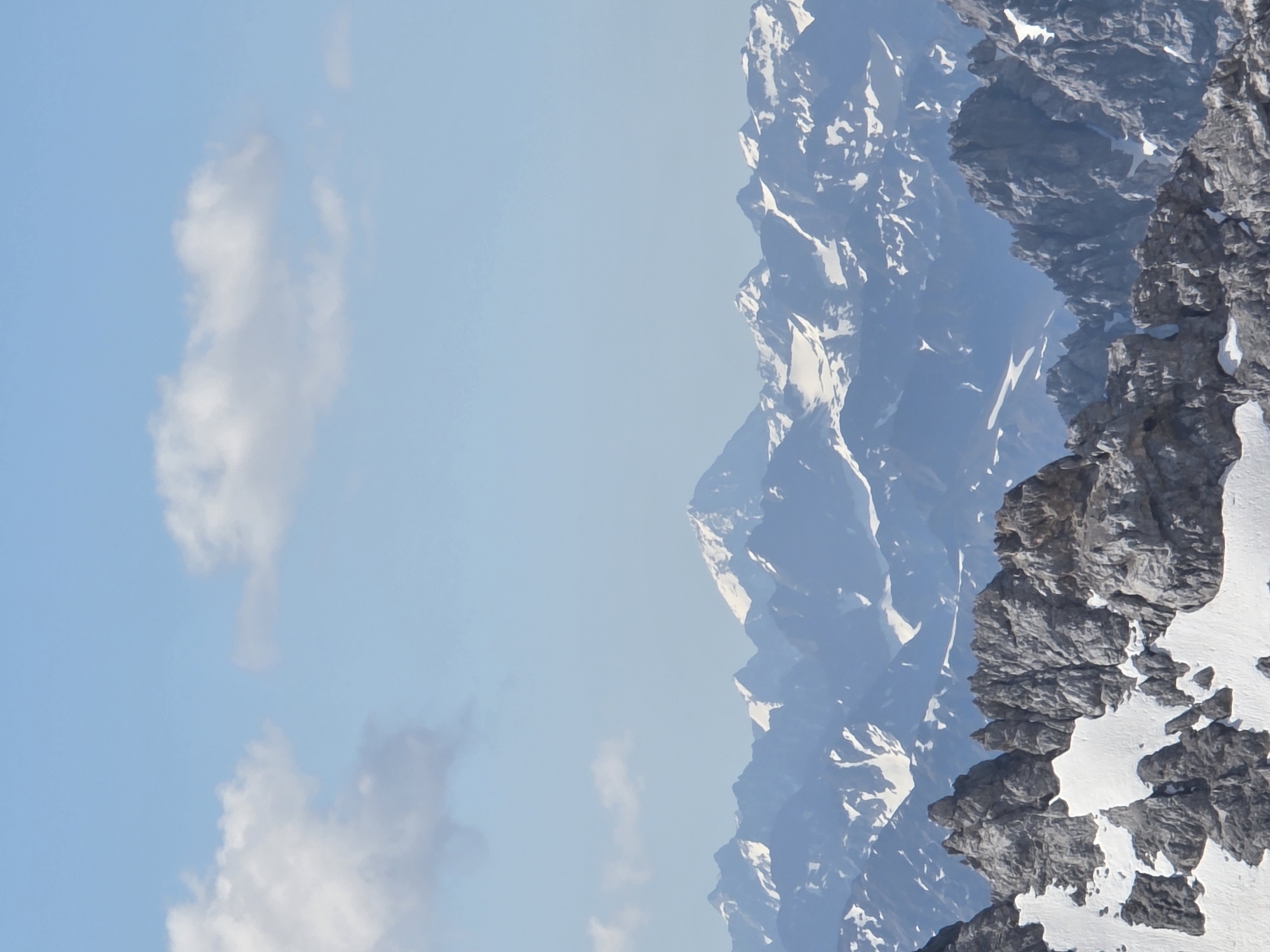 The image depicts a panoramic view of snow-capped mountains under a bright blue sky with scattered white clouds. The foreground features rugged, rocky terrain with patches of snow. In the mid-ground and background, a vast range of mountains stretches across the horizon, with some peaks appearing snow-covered and others in shadow. The overall impression is one of a majestic mountain landscape. This view is characteristic of the Bavarian Alps near Garmisch-Partenkirchen, Germany, a region renowned for its dramatic mountain scenery and popular for outdoor activities.