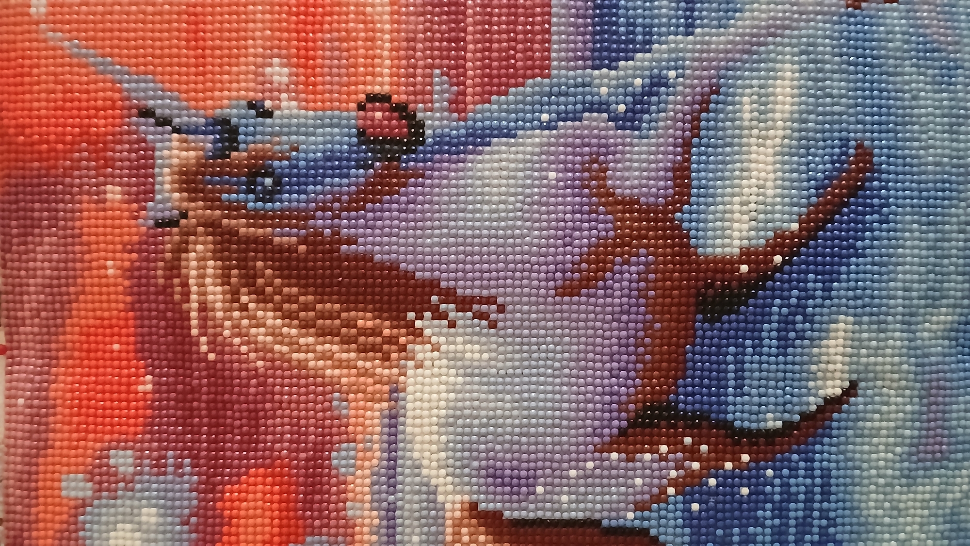 The image shows a close-up of a diamond painting featuring a deer or reindeer. The scene is rendered with small, square-shaped resin "diamonds" of varying colors, creating a pixelated effect. The color palette consists primarily of shades of red, pink, purple, blue, and brown. The deer's antlers are prominent and extend across the upper right portion of the image. The left side of the image shows the deer's head and body in tones of red and brown. The background is a gradient of colors, with red hues in the top left transitioning to cooler blues on the right. Small white diamonds are scattered throughout, possibly representing snow or highlights. The image does not provide any information about a city or location.