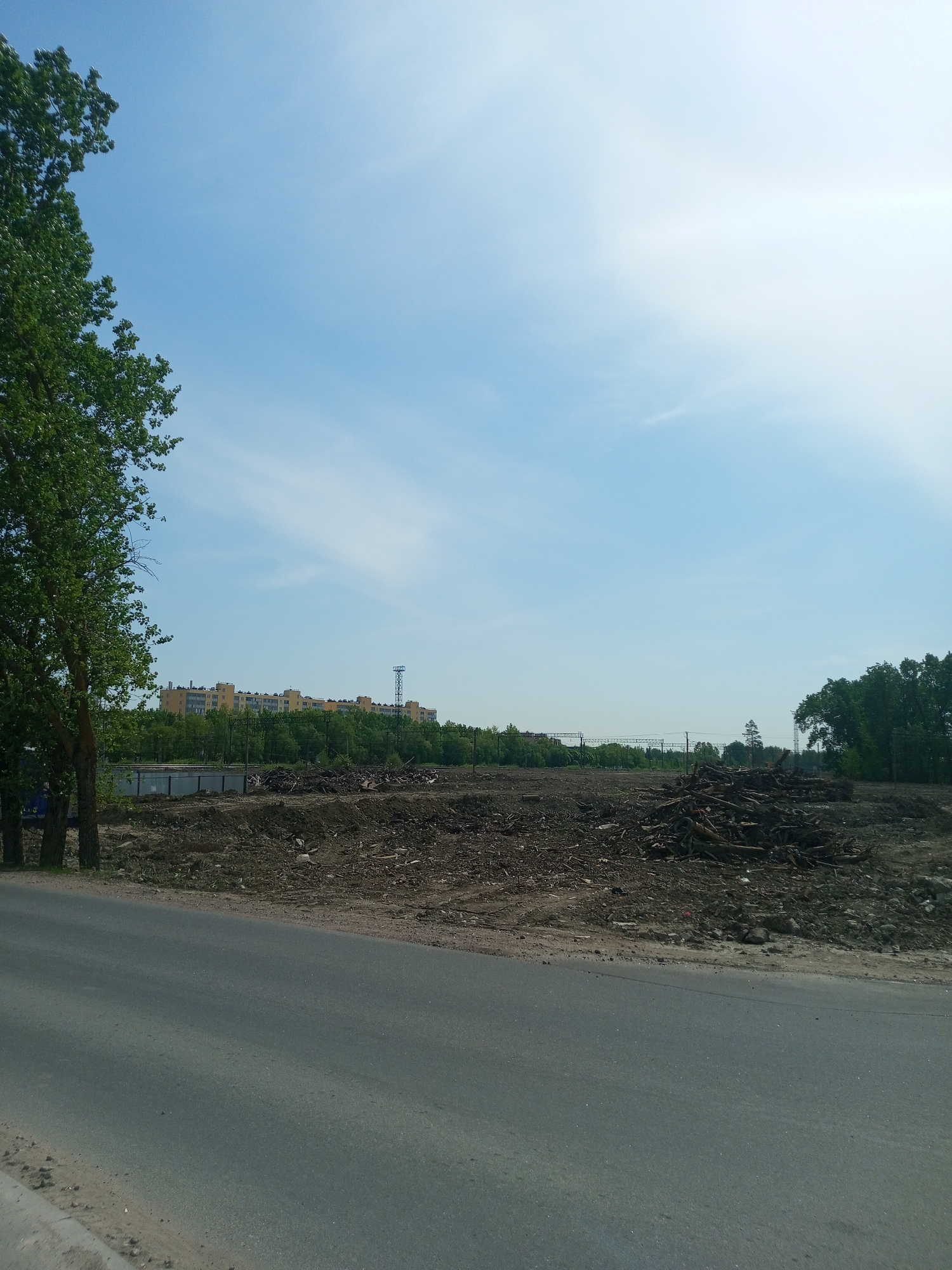 The image depicts a landscape in Kommunar, Russia. A paved road is in the foreground. Beyond the road, the scene transitions to a dirt and debris-covered area. Piles of wood debris, possibly from felled trees, are visible in the middle ground. Trees are present on both the left and right sides of the image. In the distance, buildings can be seen. The sky is blue with scattered clouds.