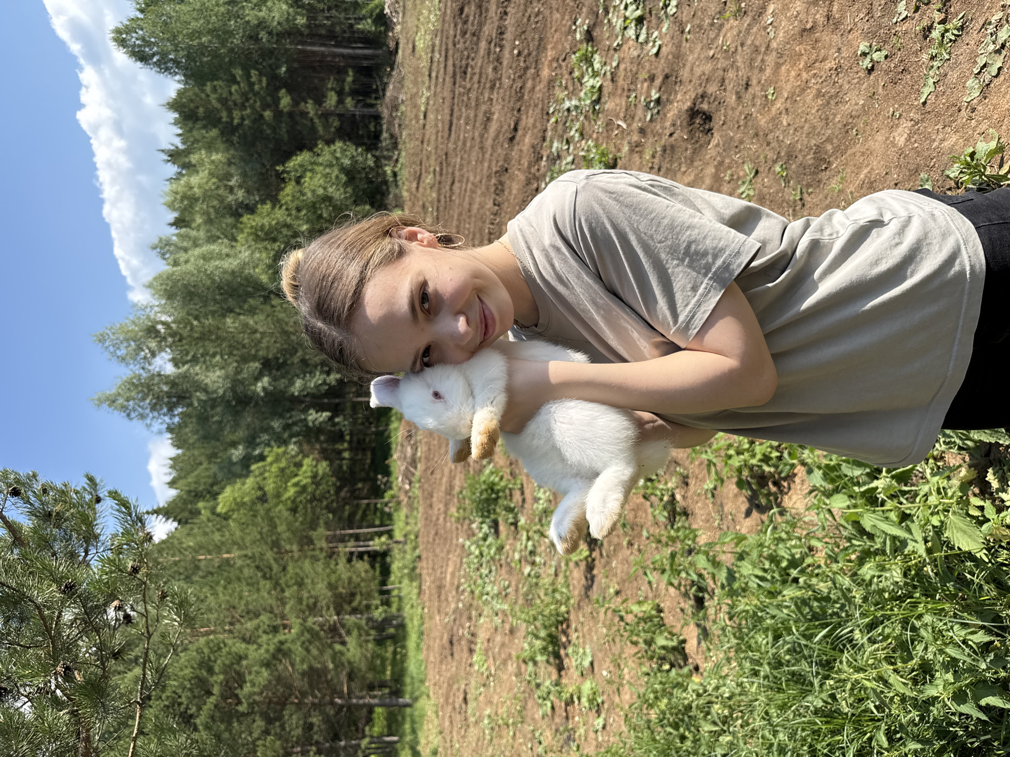 A young woman, appearing to be in her late teens or early twenties, is holding a white rabbit against her shoulder. She has light brown hair pulled back in a bun and is wearing a grey t-shirt and dark pants. She is smiling and looking at the camera. The background suggests a rural or natural setting, with trees, a field, and a dirt path visible. The overall impression is one of a serene, outdoor moment, possibly in a rural area. The specific location in the city of Golyan, Russia, is not discernible from the image itself; this information would need to be provided externally.